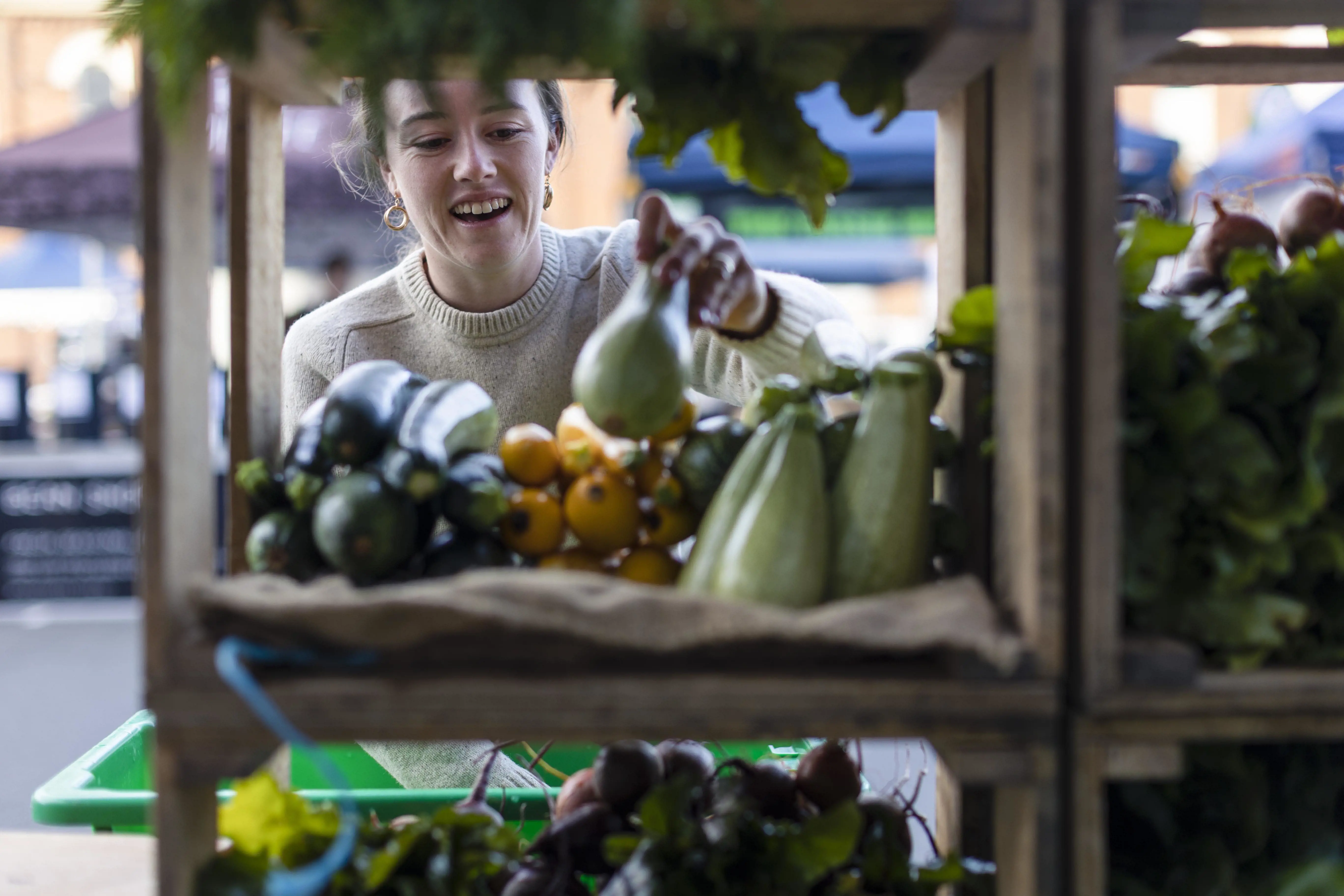 A young woman arranges fresh vegetables in square wooden shelves in a market space. 