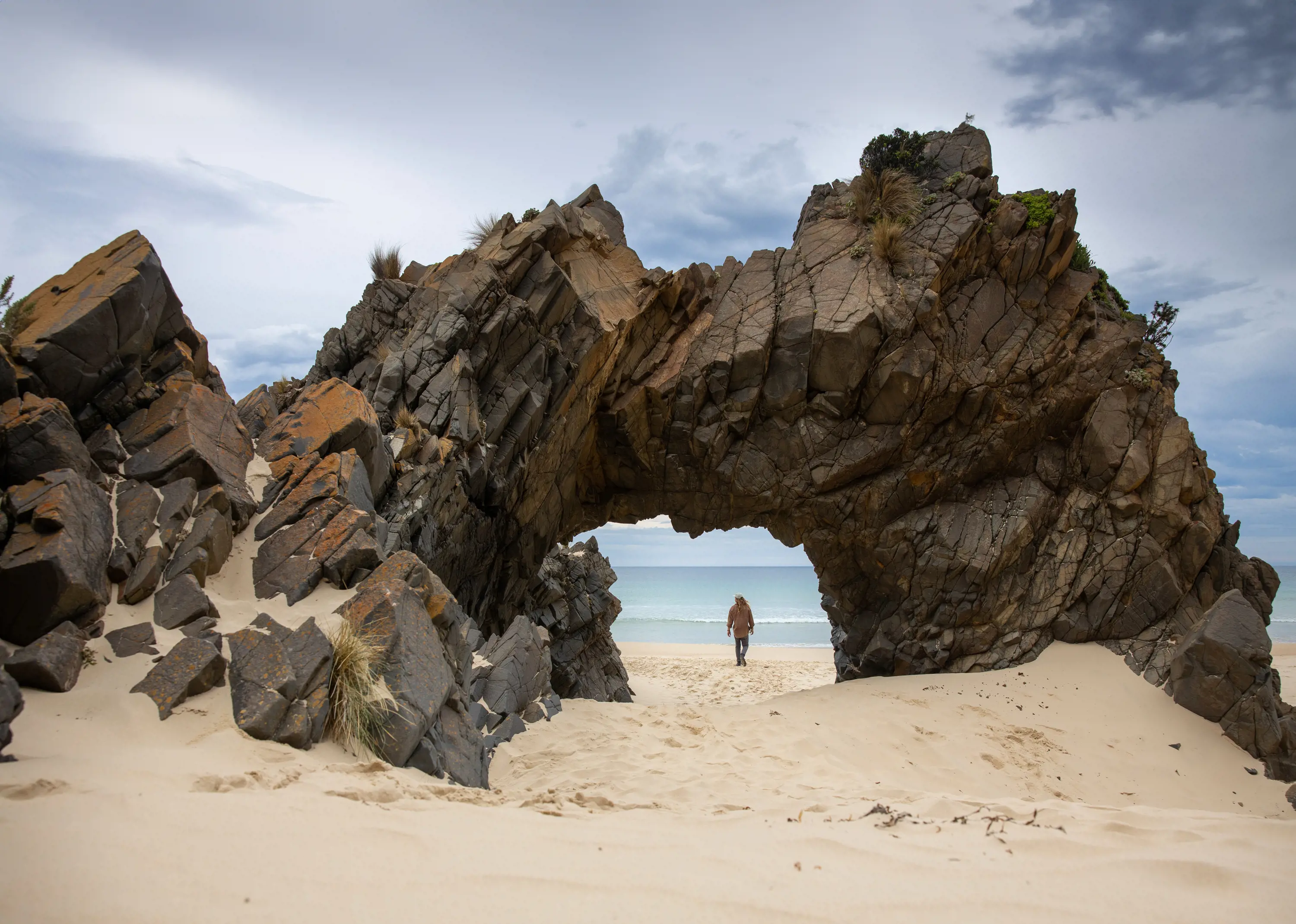 A man walks along the sand, seen through the arch of a large rock formation jutting out of sand in the foreground.