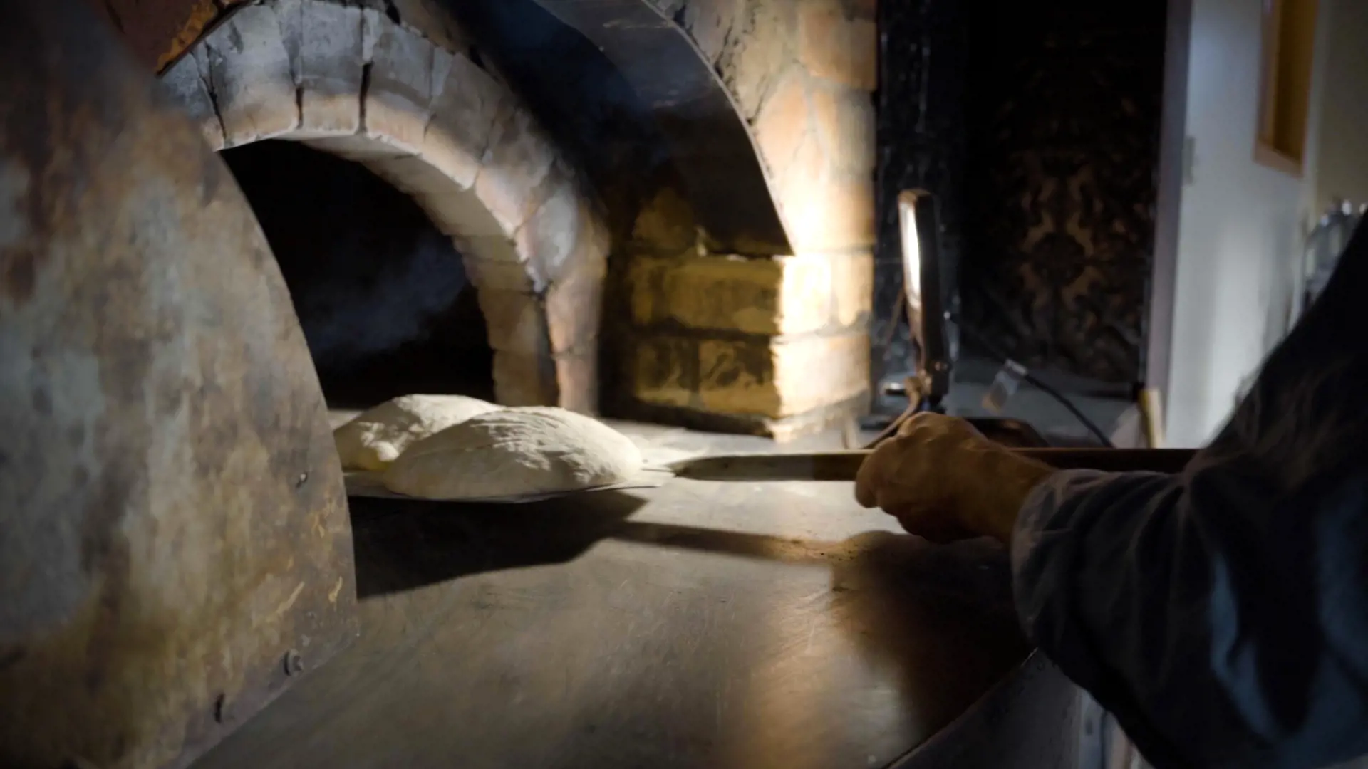 A man carefully places bread dough into the arched opening of a large brick wood-fired oven.