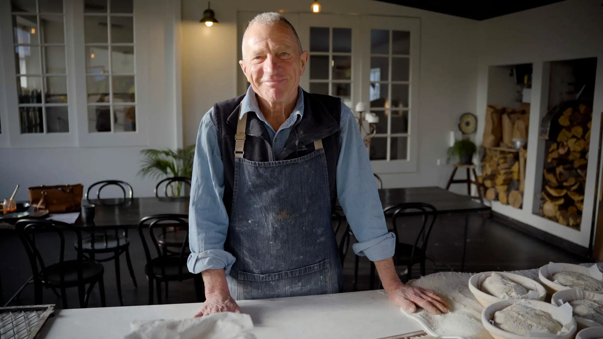 A older man wearing a denim apron and a blue shirt stands behind a large table covered in bread dough.