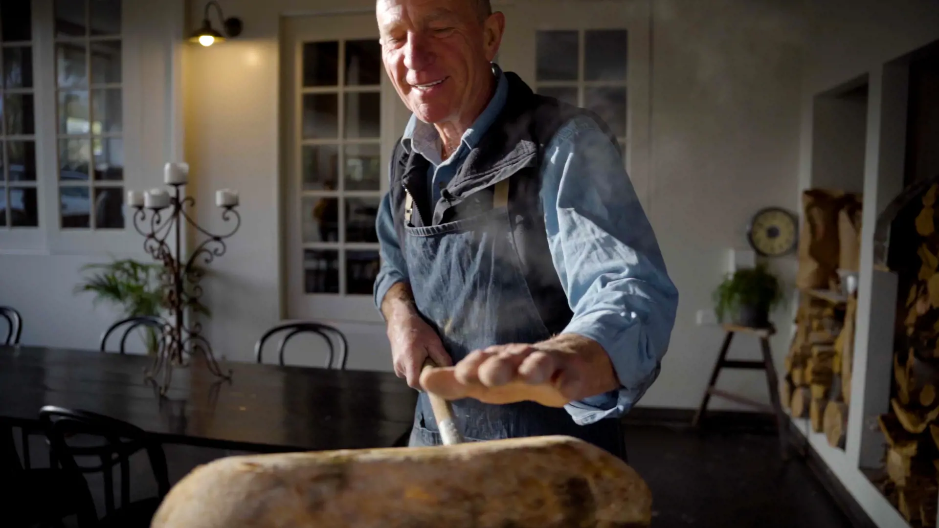 An older man wearing a denim apron and a blue shirt pulls a freshly cooked loaf of handmade bread from the oven.