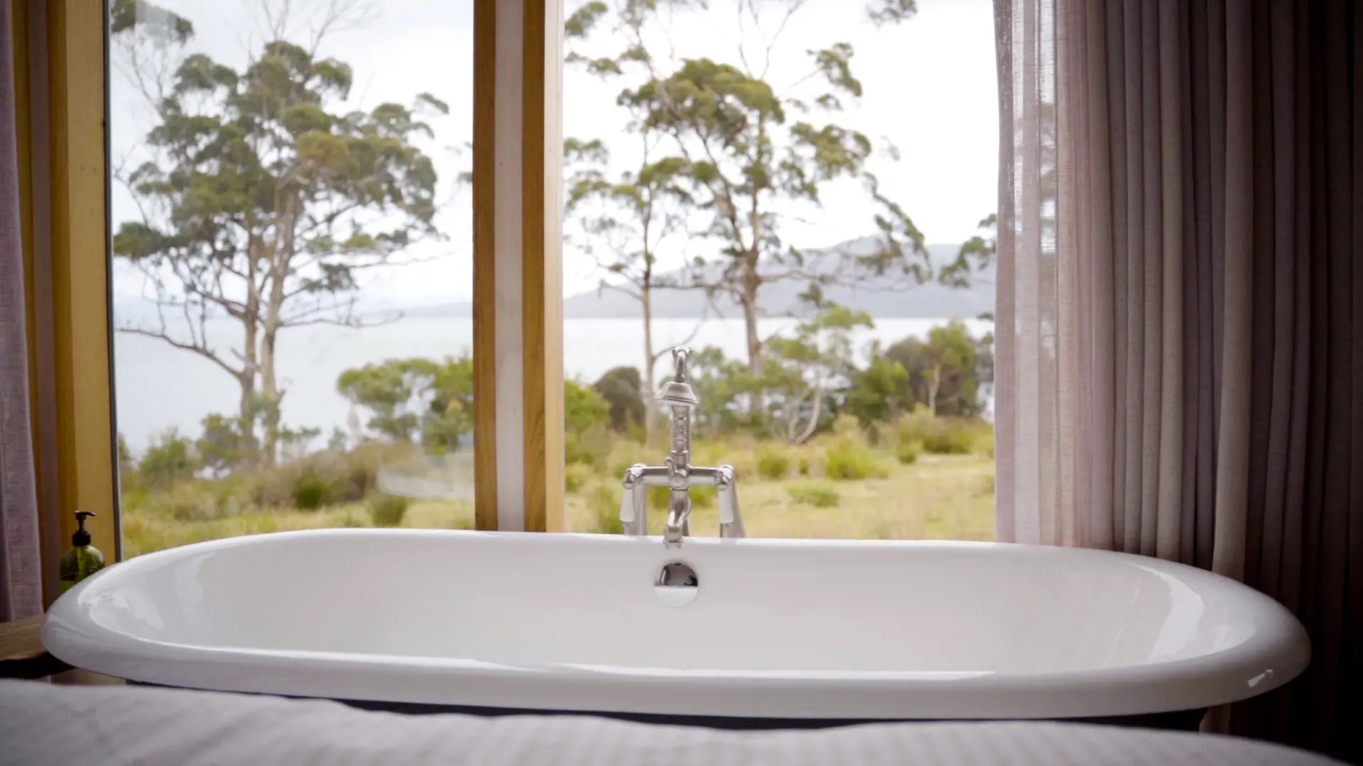 A large bath sits next to a window looking out through bushland to the sea.