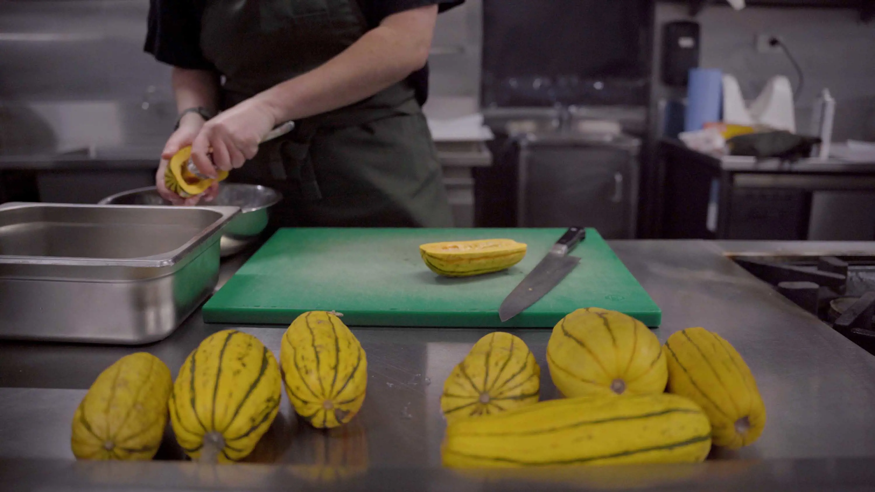 7 Delicata squash sit on a stainless steel cooking benchtop in the foreground. A chef scoops out the seeds of the yellow squash using a spoon.