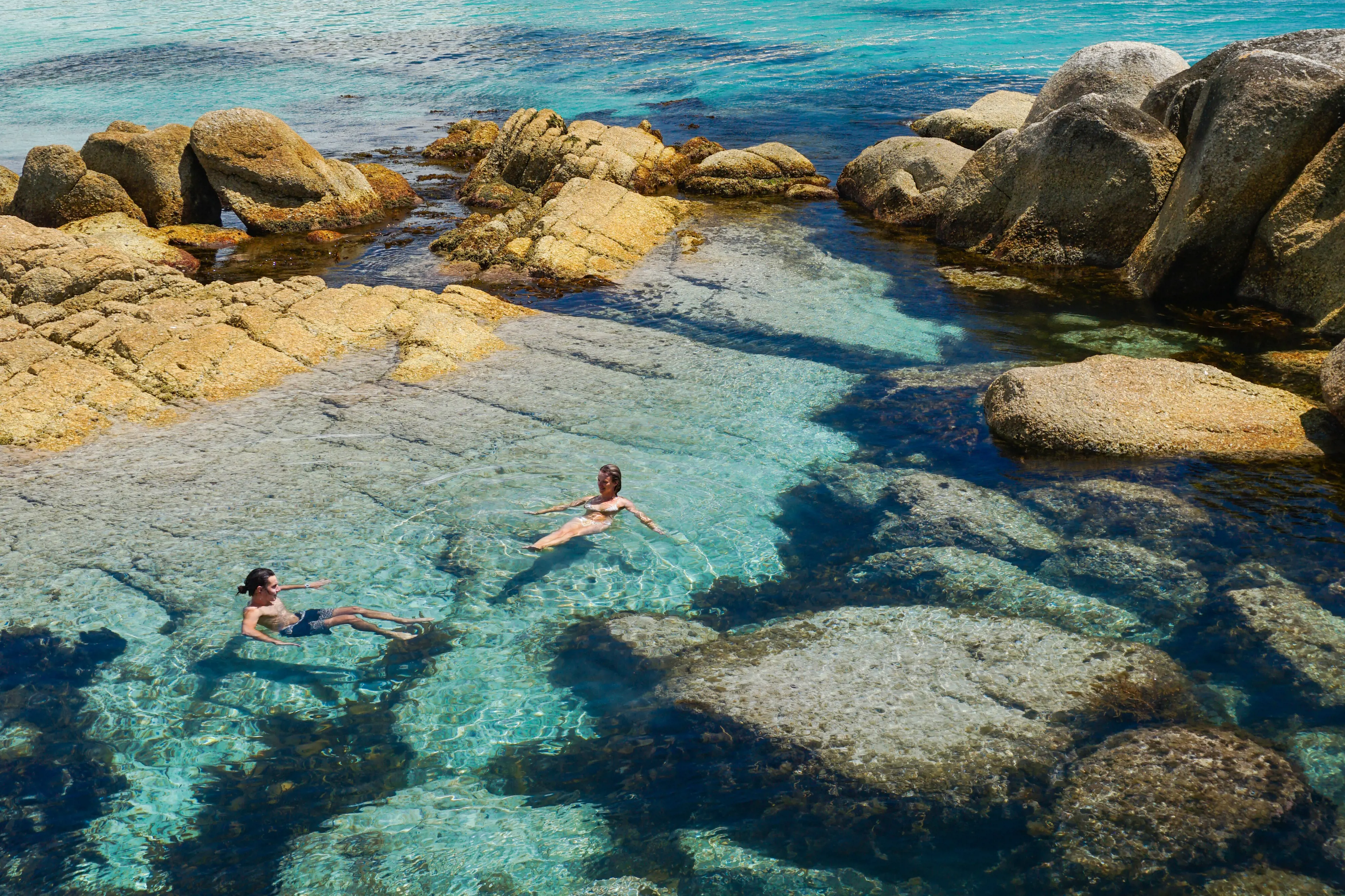 Swimmers float in the light blue waters of a rockpool on a sunny sunny day.