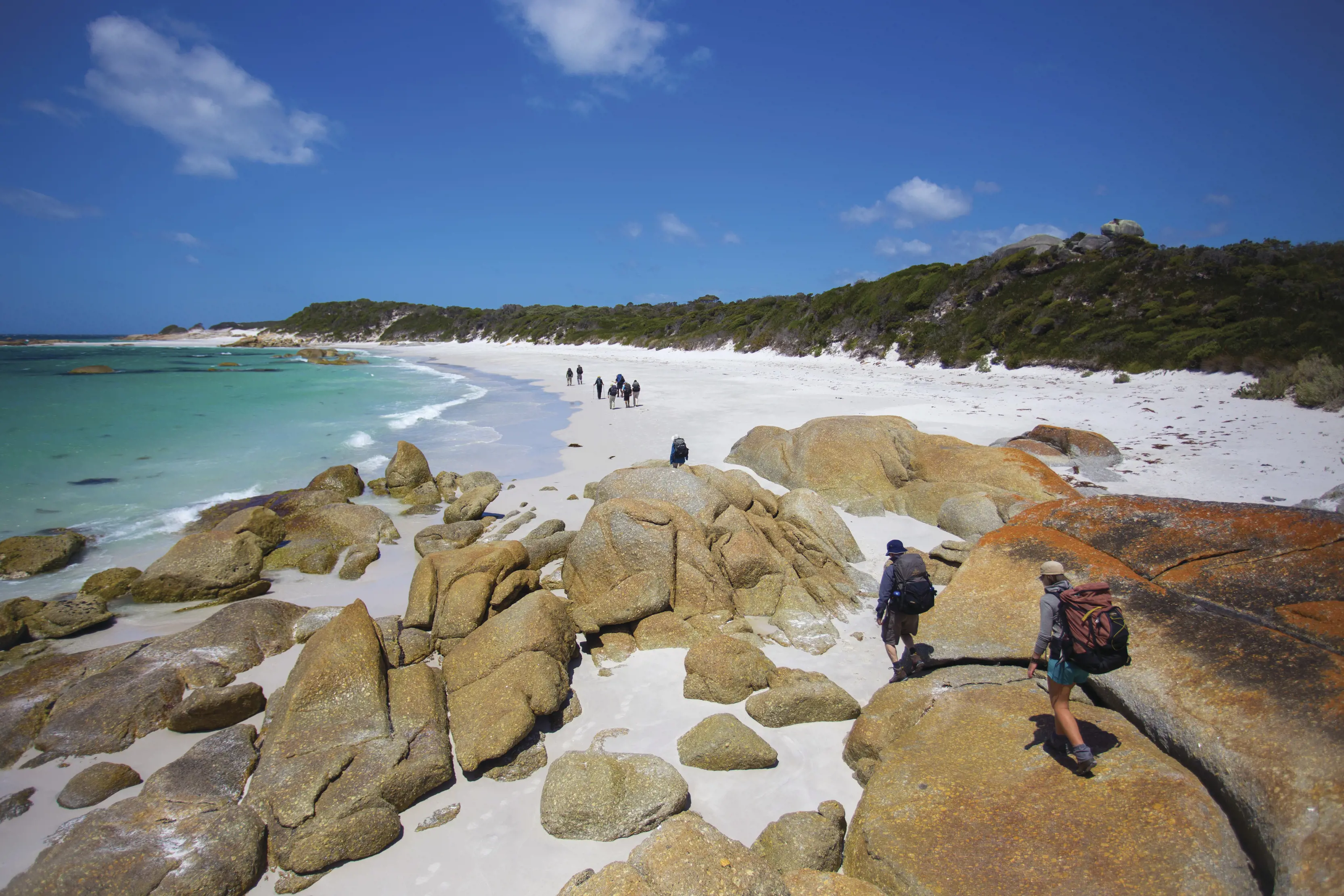 In the foreground, hikers climb down the rocks at Bay of Fires Lodge Walk. In the distance, there are more walkers on the beach.