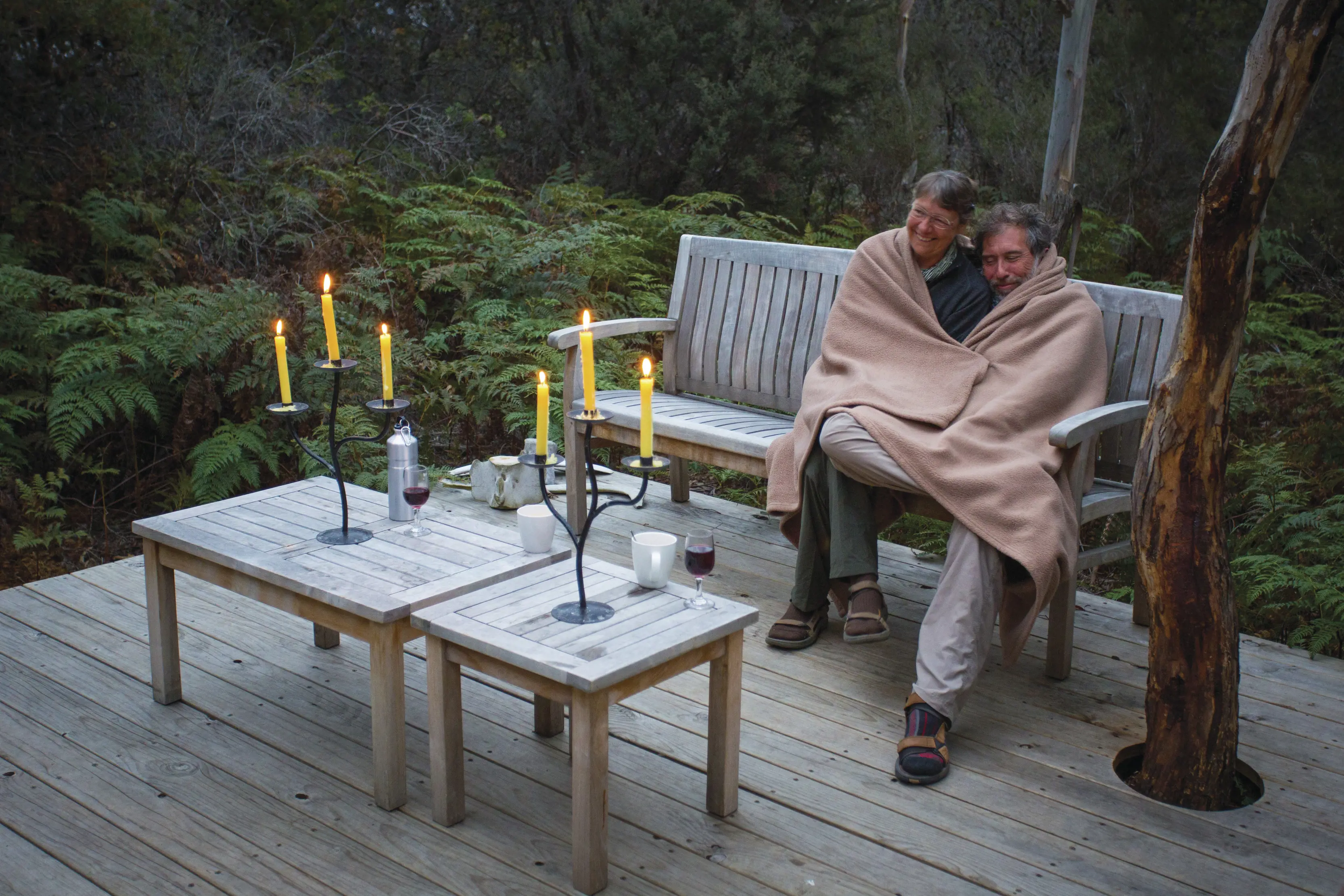 A couple wrap themselves in a shared blanket whilst sat at a table lit table surrounded by the rainforest on the Maria Island Walk,