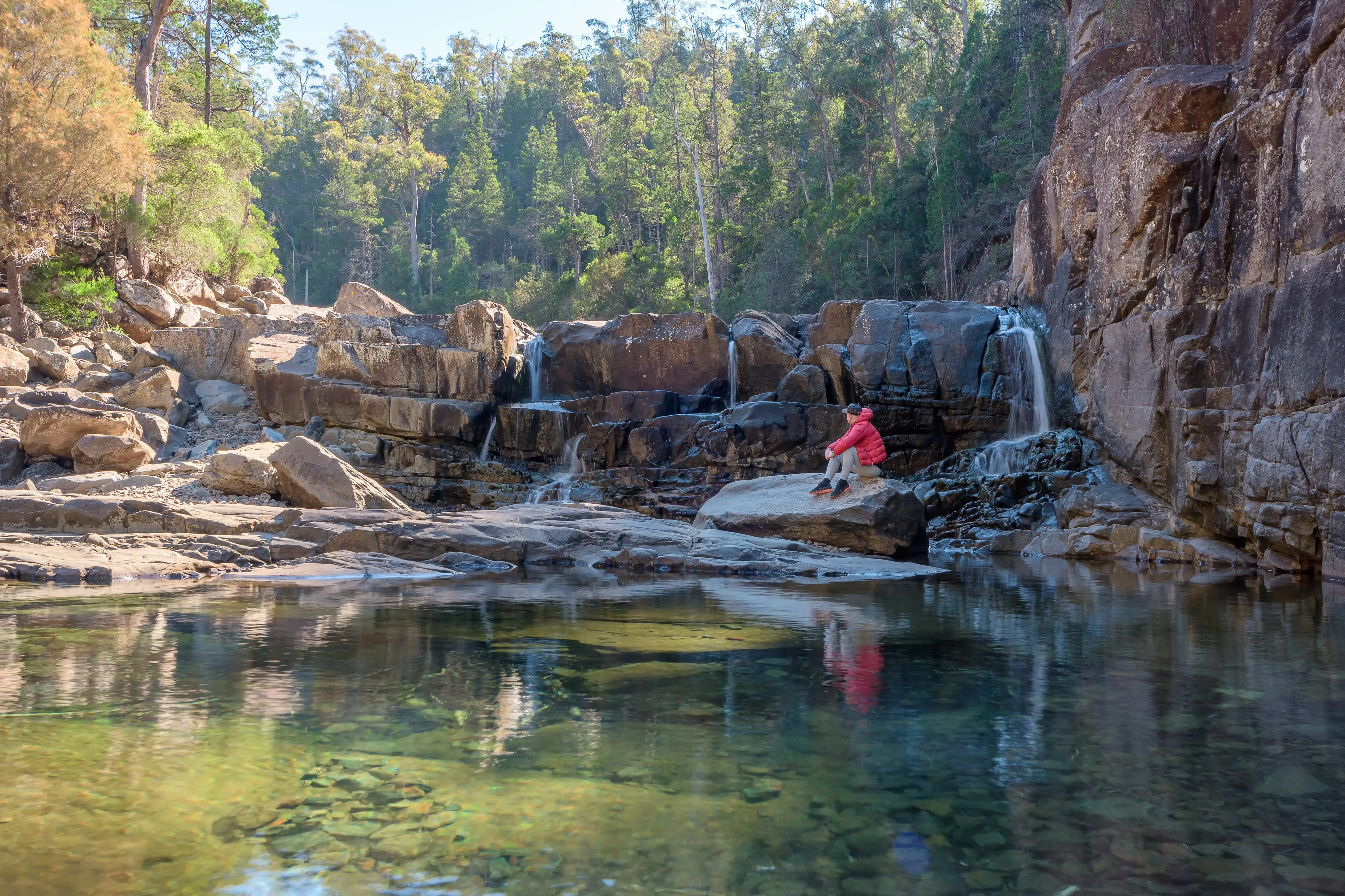 In the foreground, a man sits on the rocks by a tranquil pool at Apsley Gorge. There is rainforset in the back drop.