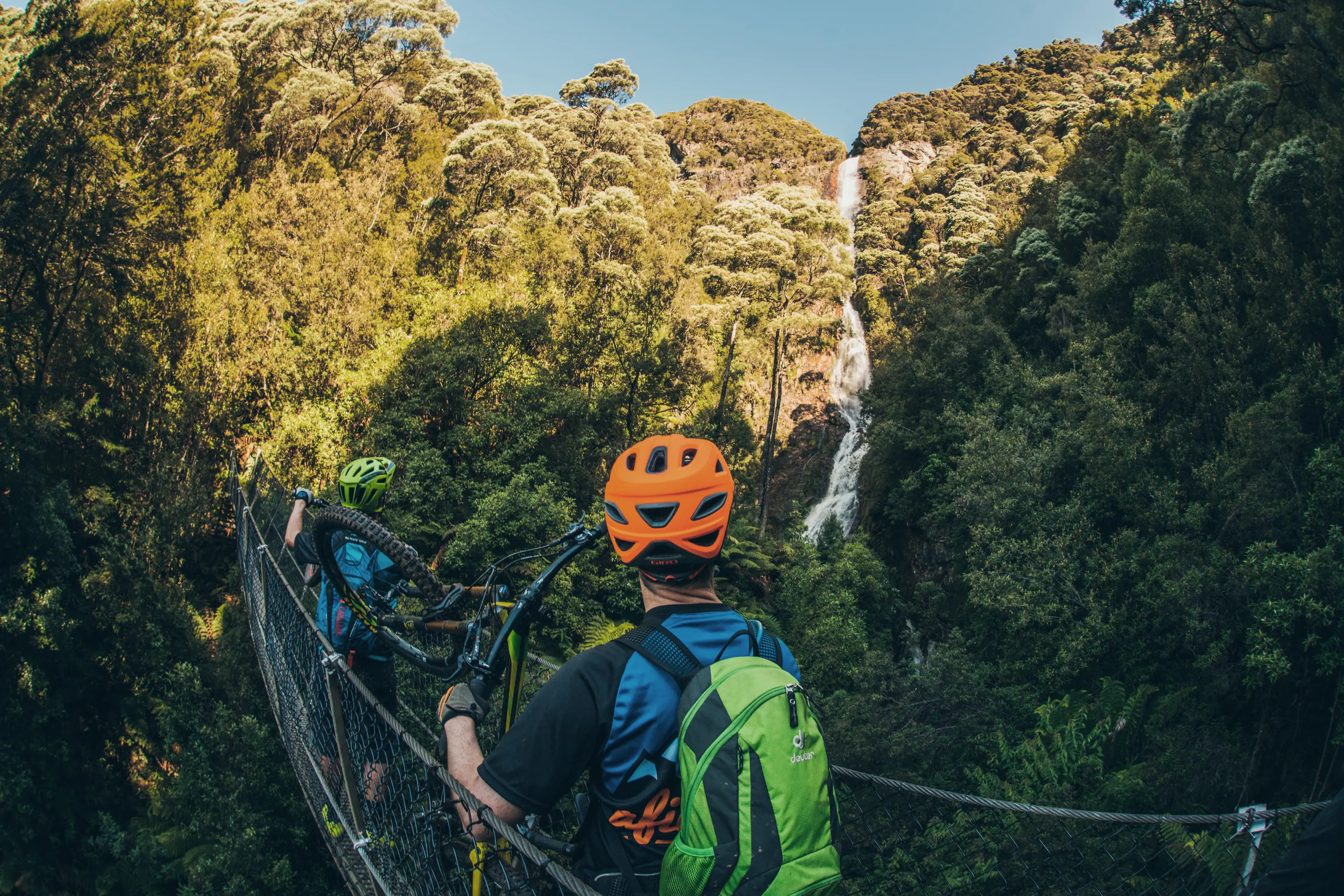 A man wearing mountain bike gear stands on a narrow suspension bridge and looks out over forest to Monetzuma Falls.