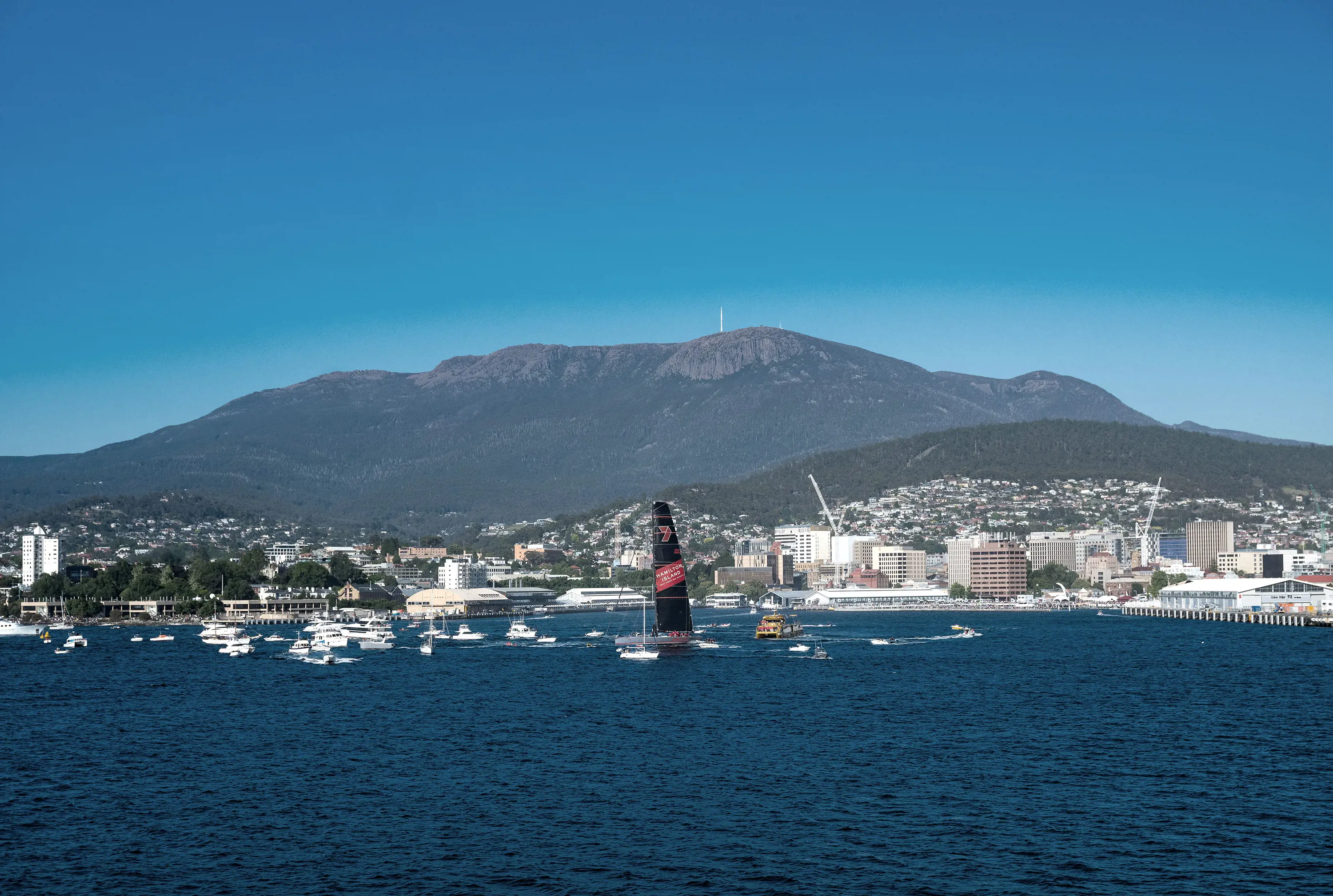 A large racing yacht and many small cruising yachts circle around in the waters around the Hobart docks.