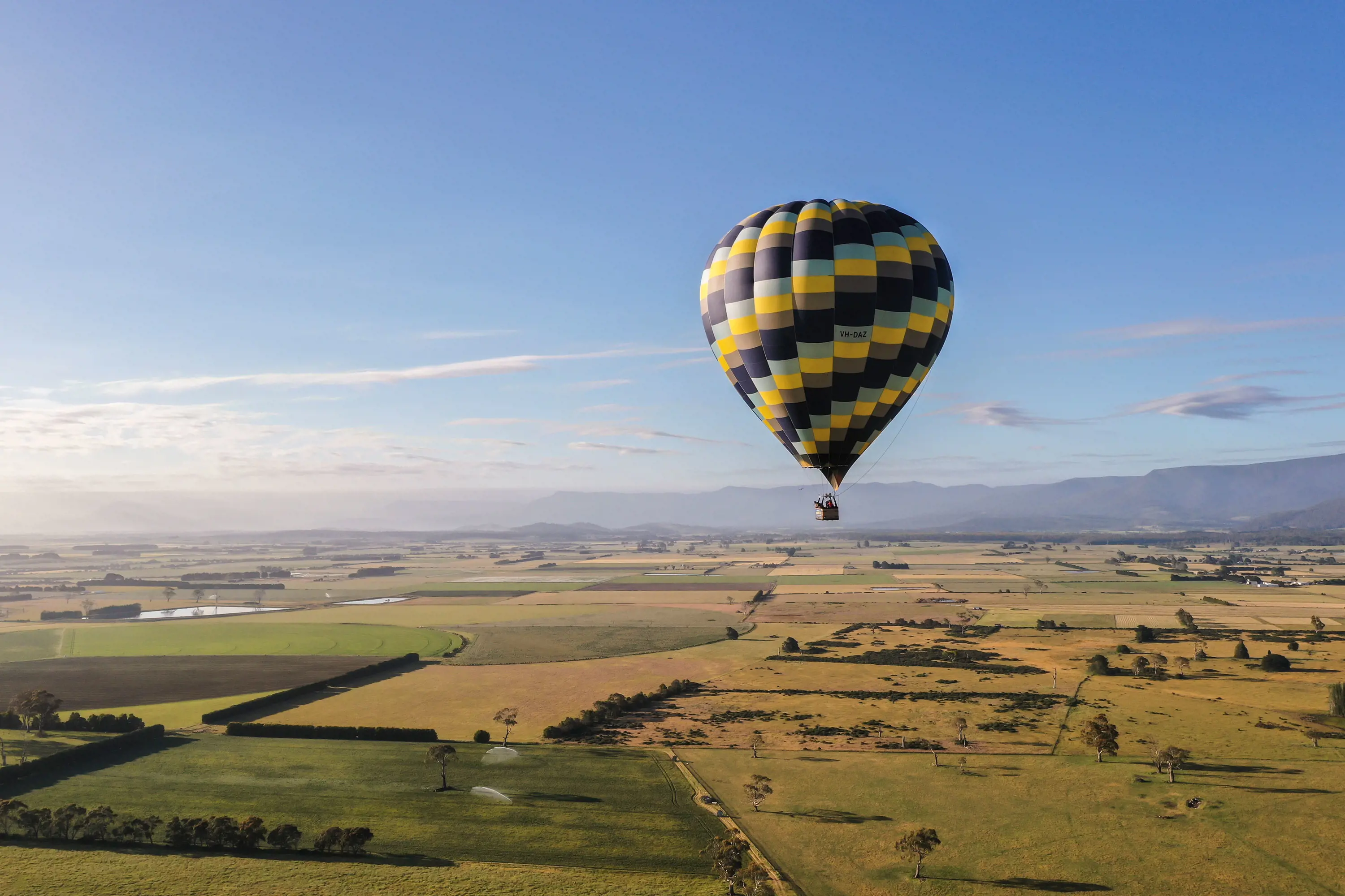A large yellow and purple patterned air balloon sails peacefully over farmlands on a clear, sunny day.