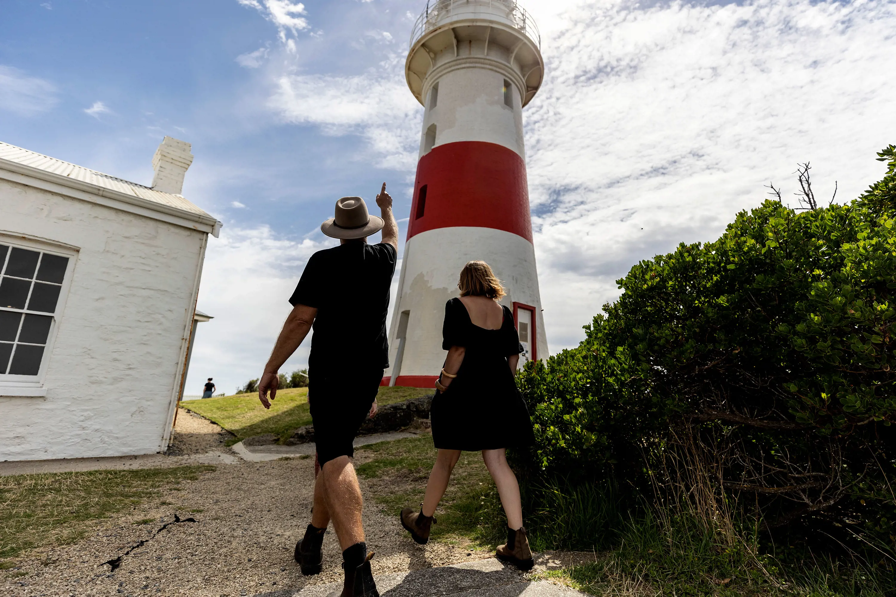 A guide and a tourist walk along a gravel path below a tall, red and white lighthouse.