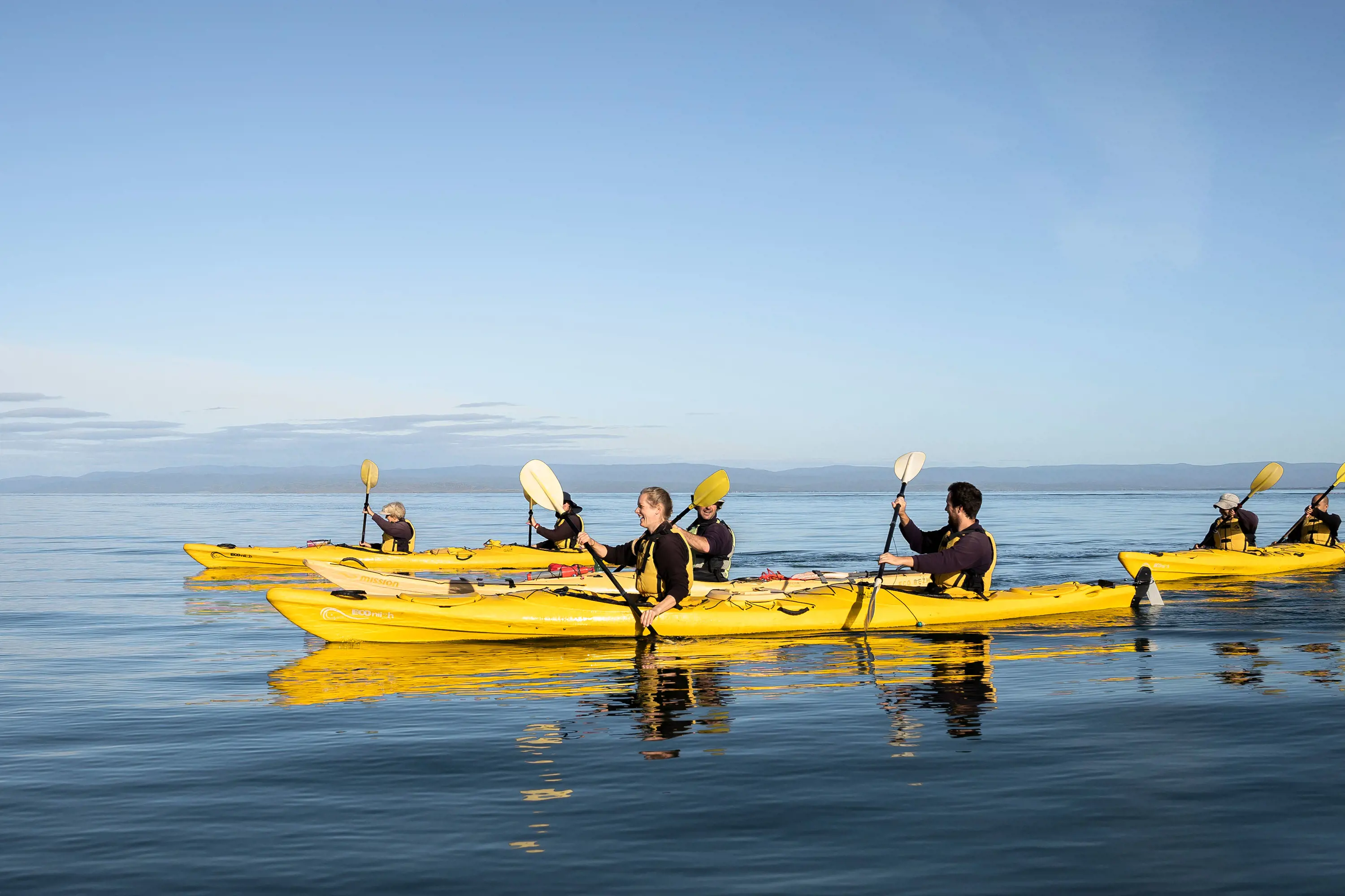 A group of 7 kayakers in bright yellow kayaks paddle across calm, dark blue waters.