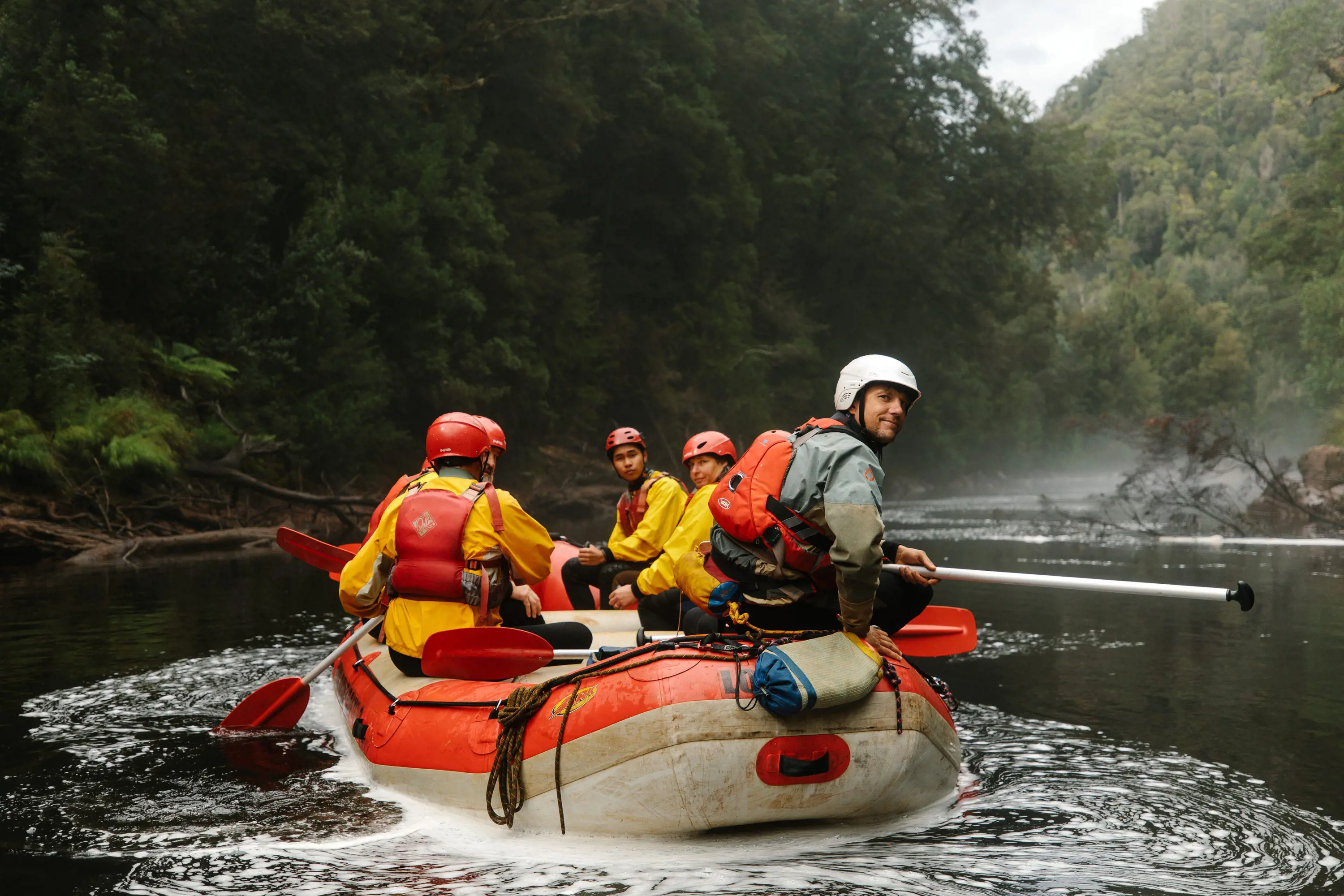 A group wearing life jackets and safety helmets sit in a large raft and paddle along calm waters in a river, passing through dense forest.