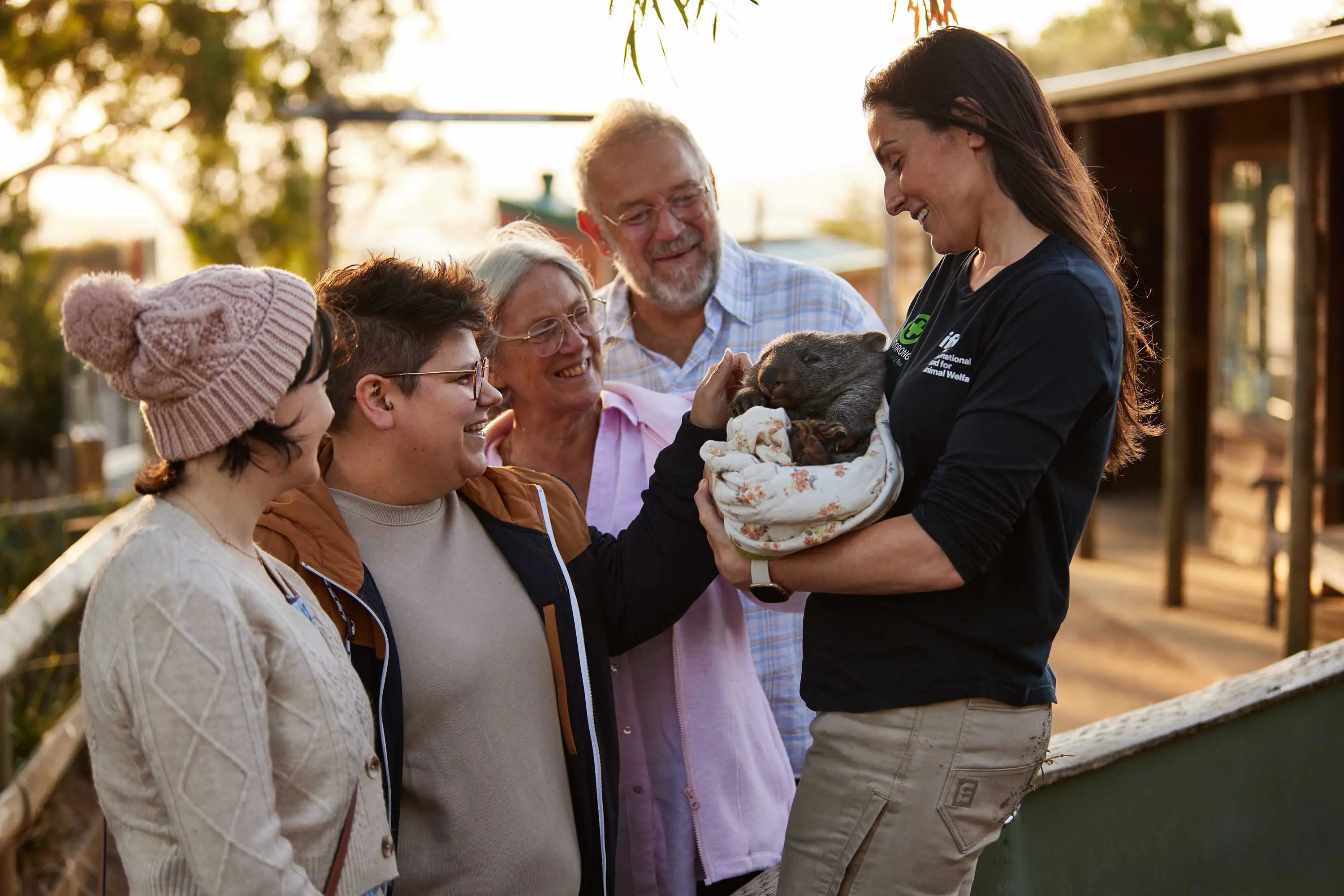 A wildlife handler cradles a baby wombat wrapped in a white blanket and shows a small group of smiling people.