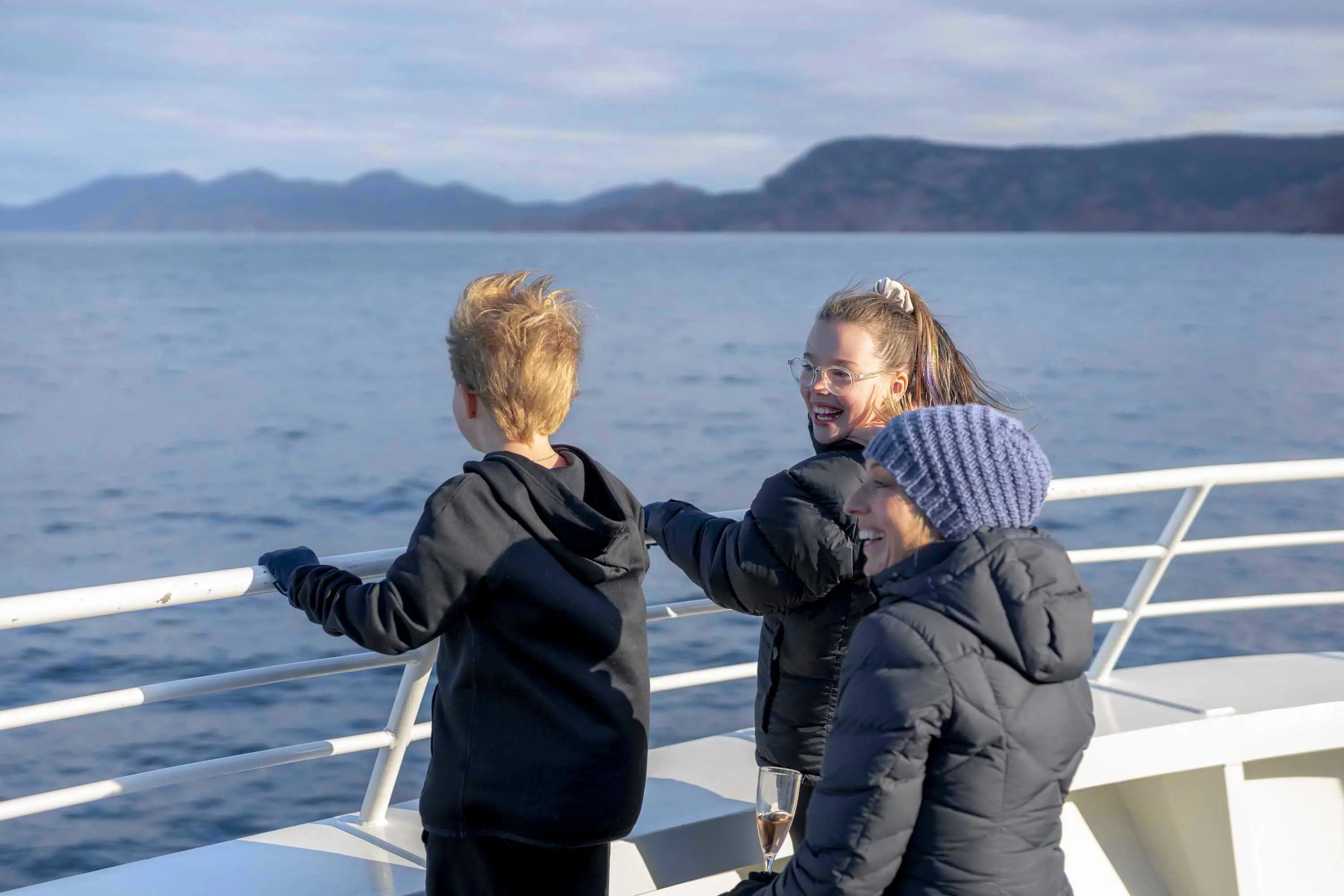 A mother and her young children stand on the deck of a cruise boat and look out over the blue water. Red rocked mountains are in the distance.