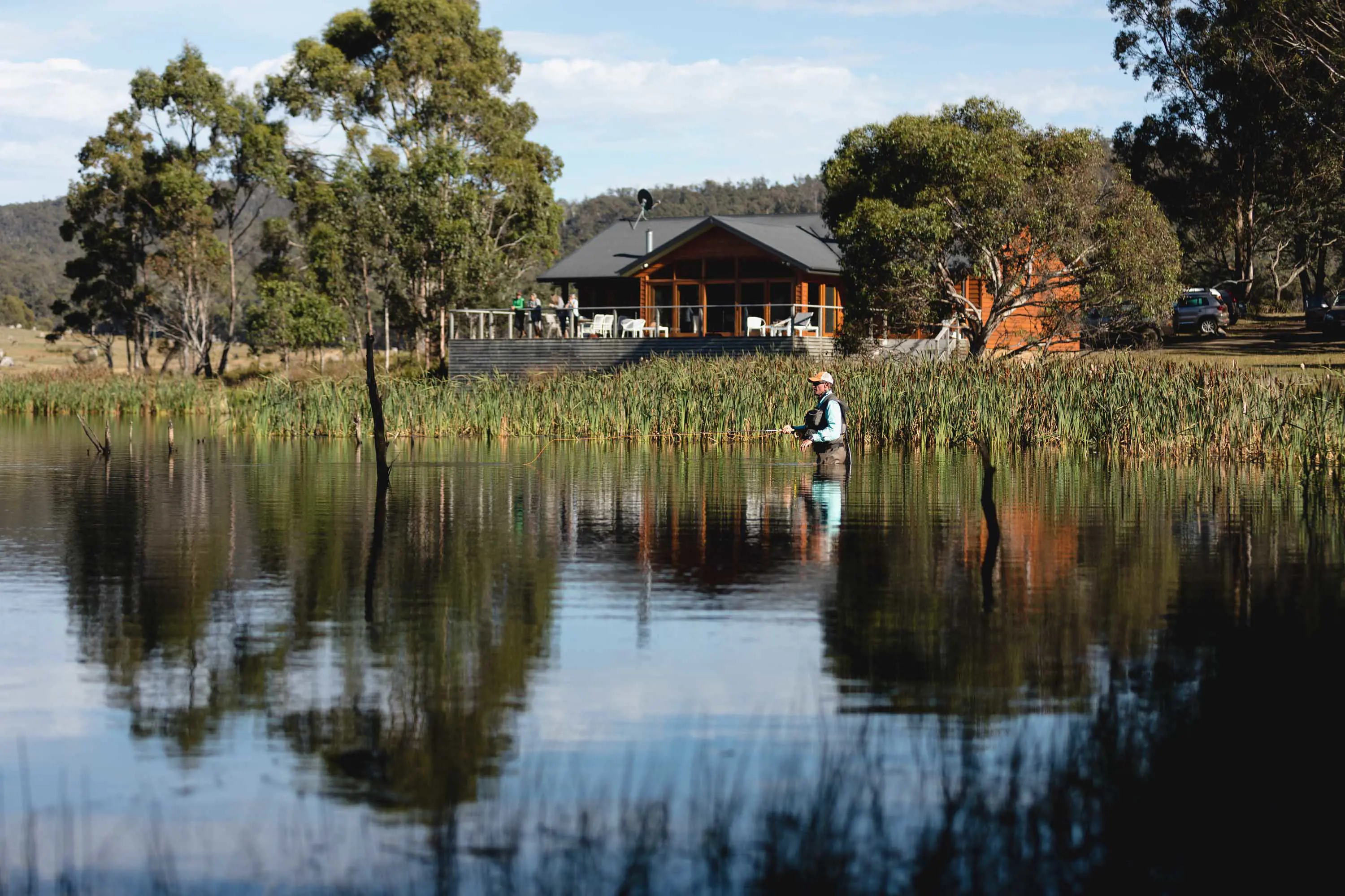A man stands up to his waist in stilll waters fishing, with reeds and wooden house in the background.