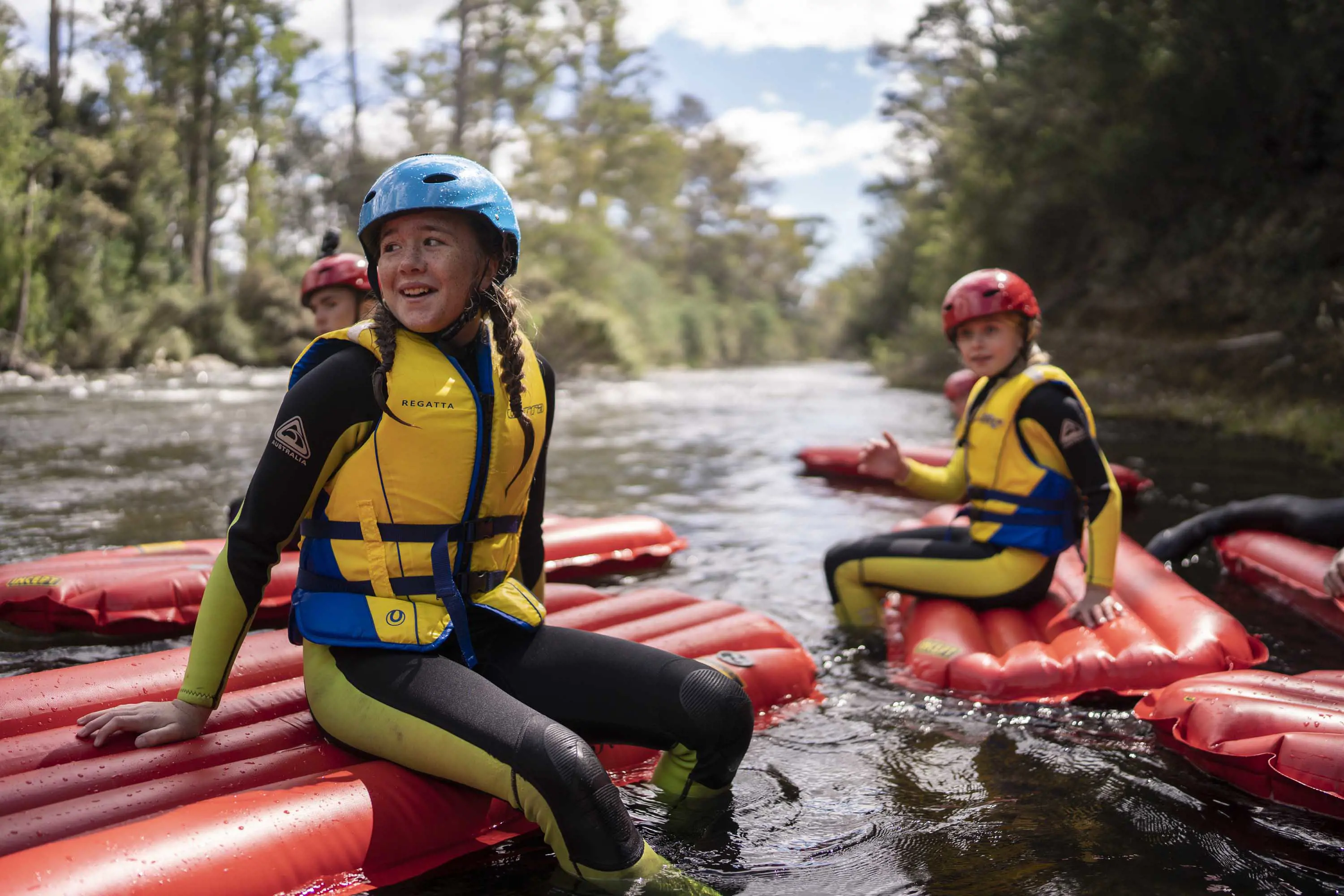 Children in floatation vests, wetsuits and protective head wear sit on red, inflatable plastic rafts in a river with a mild current.
