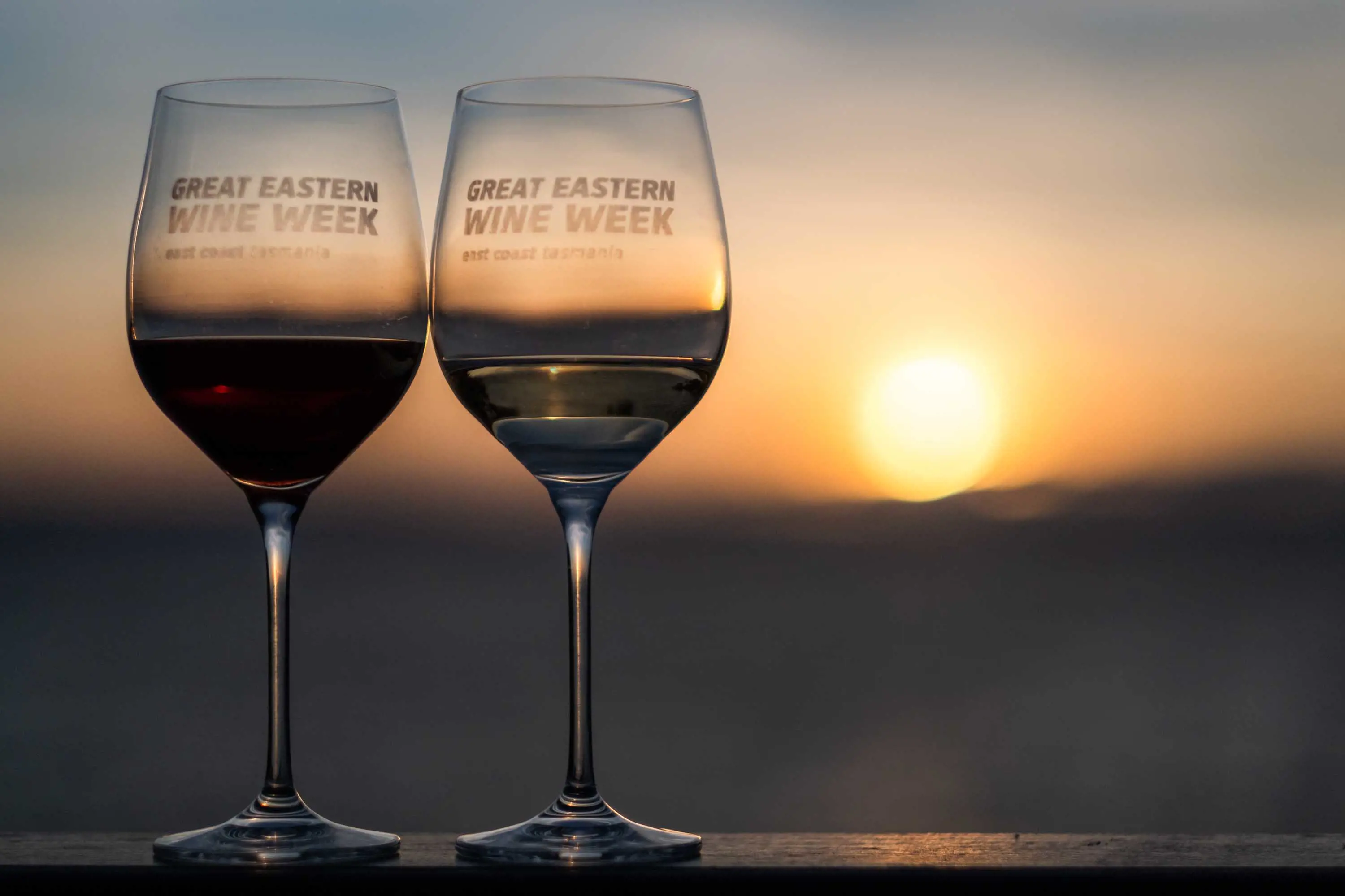 Two wine glasses filled with white wine sit on a bench with the sun setting in the distance.