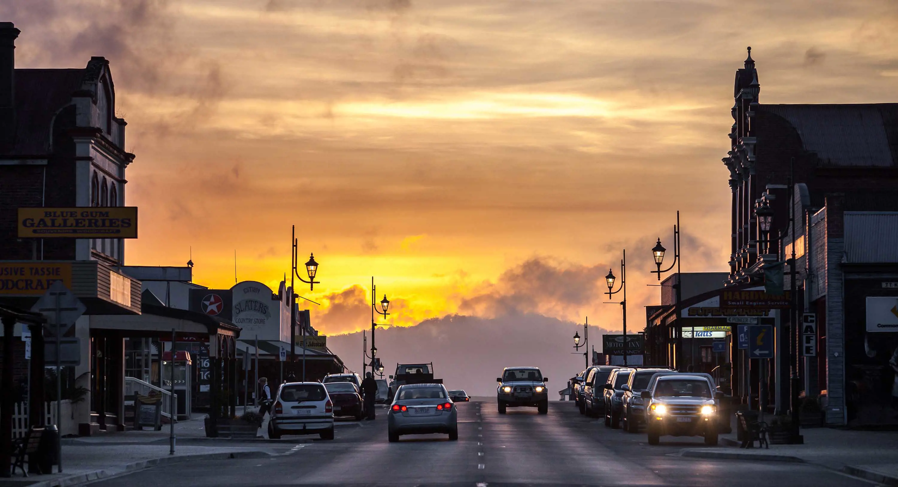 A wide street with cars travelling in both directions lined with old buildings at sunset.