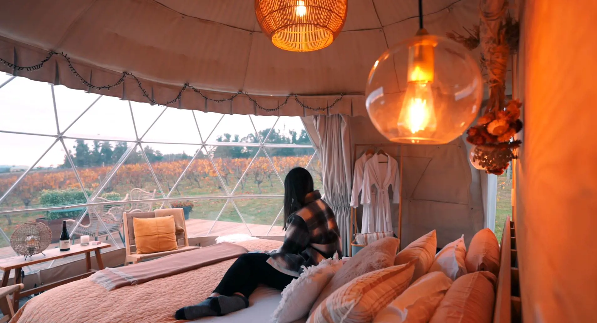 A woman sits on a large bed inside a dome-shaped tent with windows overlooking a vineyard.
