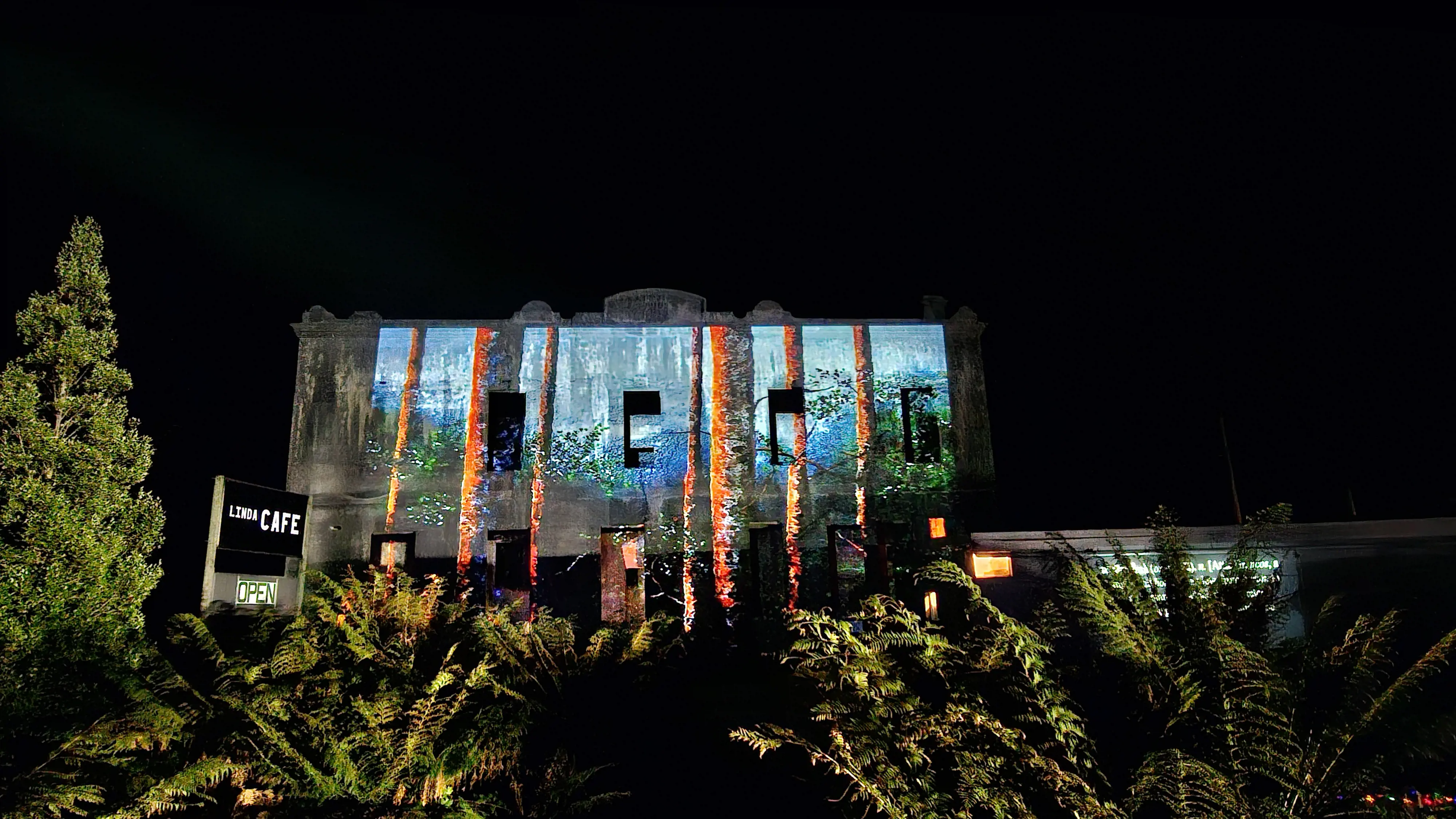 A bright projection of tall pine trees in a forest is displayed on the front facade of the Royal Hotel near the cafe.