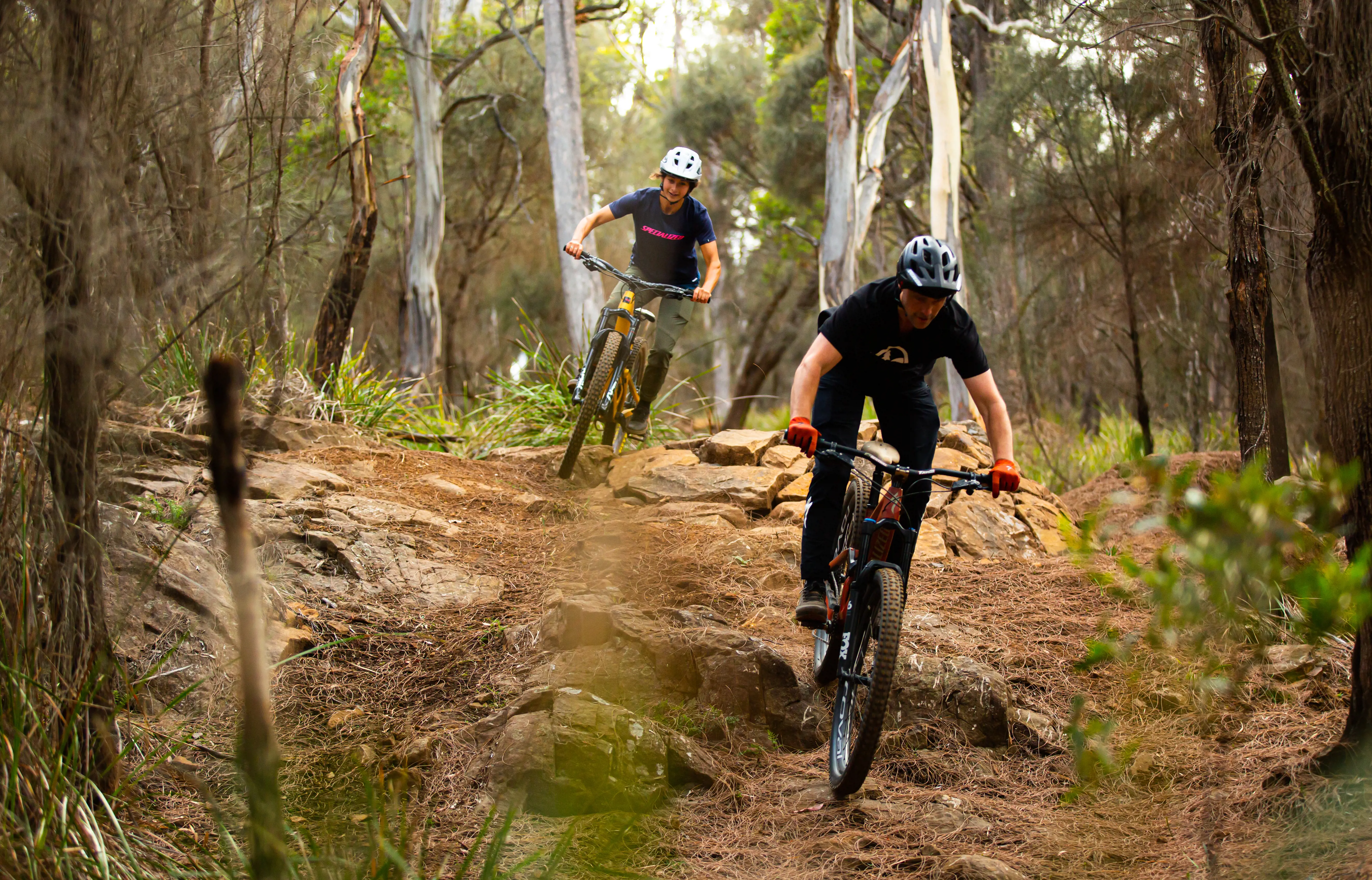 A pair of mountain bike riders negotiate rocks on a dusty trail through dense scrub.