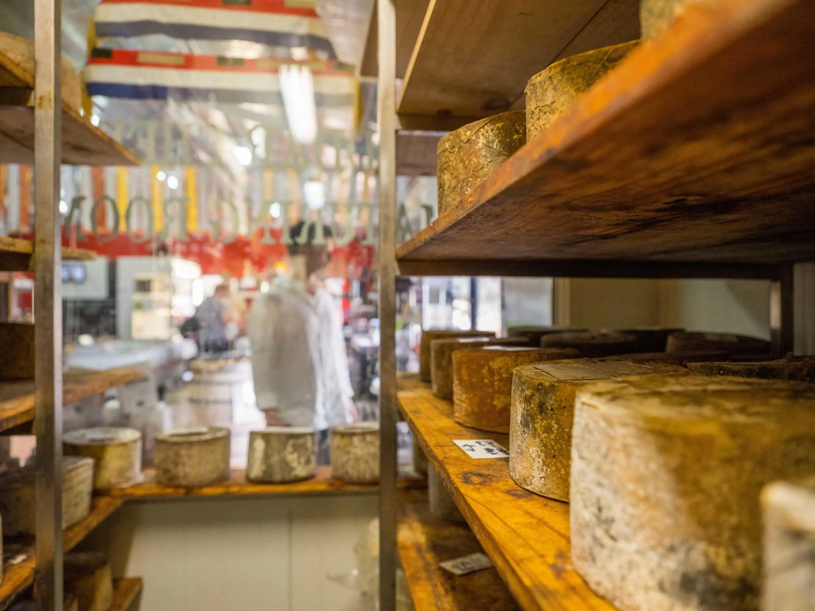 Handmade cheese is placed on wooden shelves in a refrigerated room with a window out onto a factory floor.