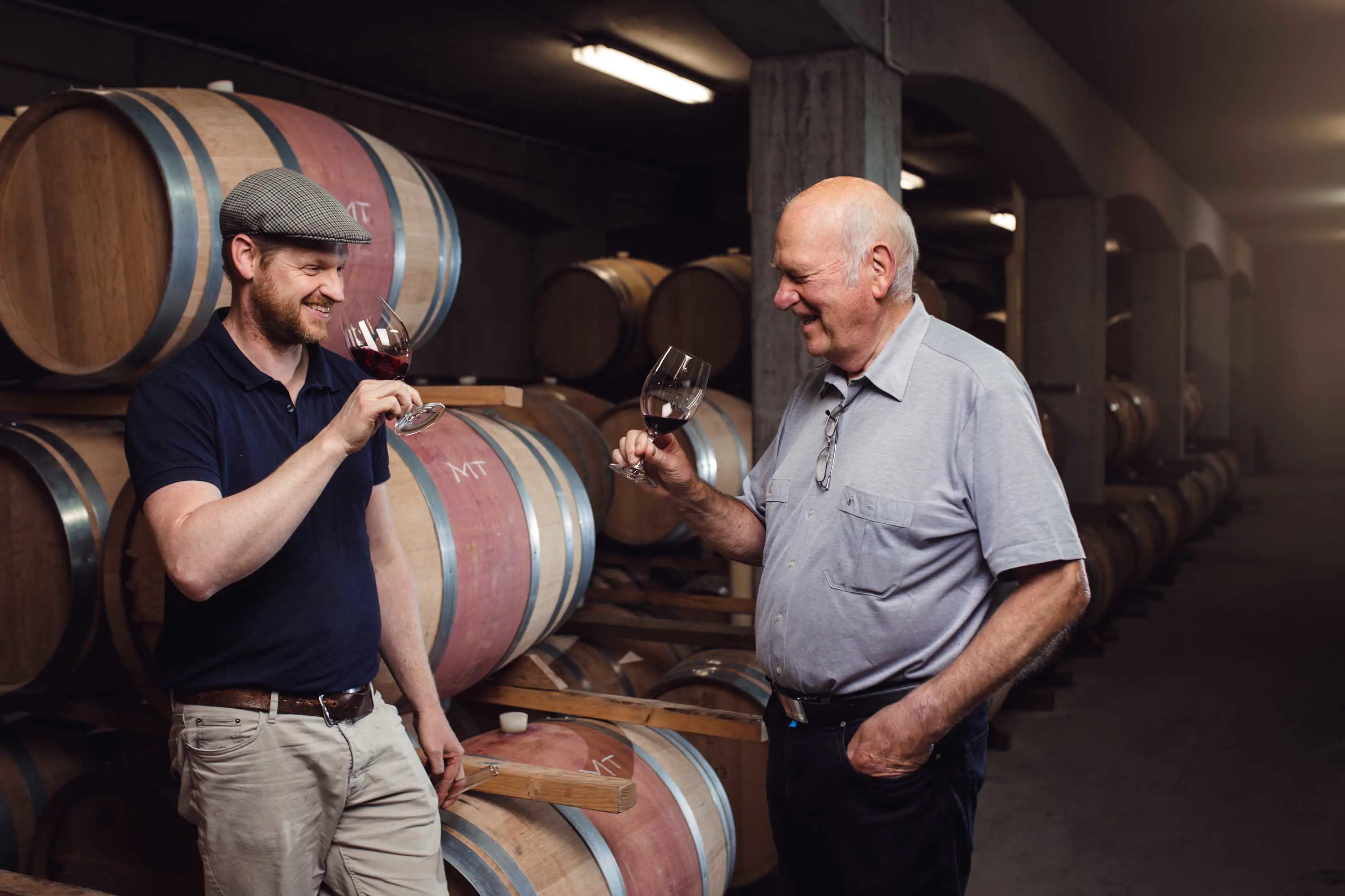 A young man wearing a flat cap and an older man in a buttoned shirt drink wine, standing in front of barrels stacked up in a concrete warehouse.