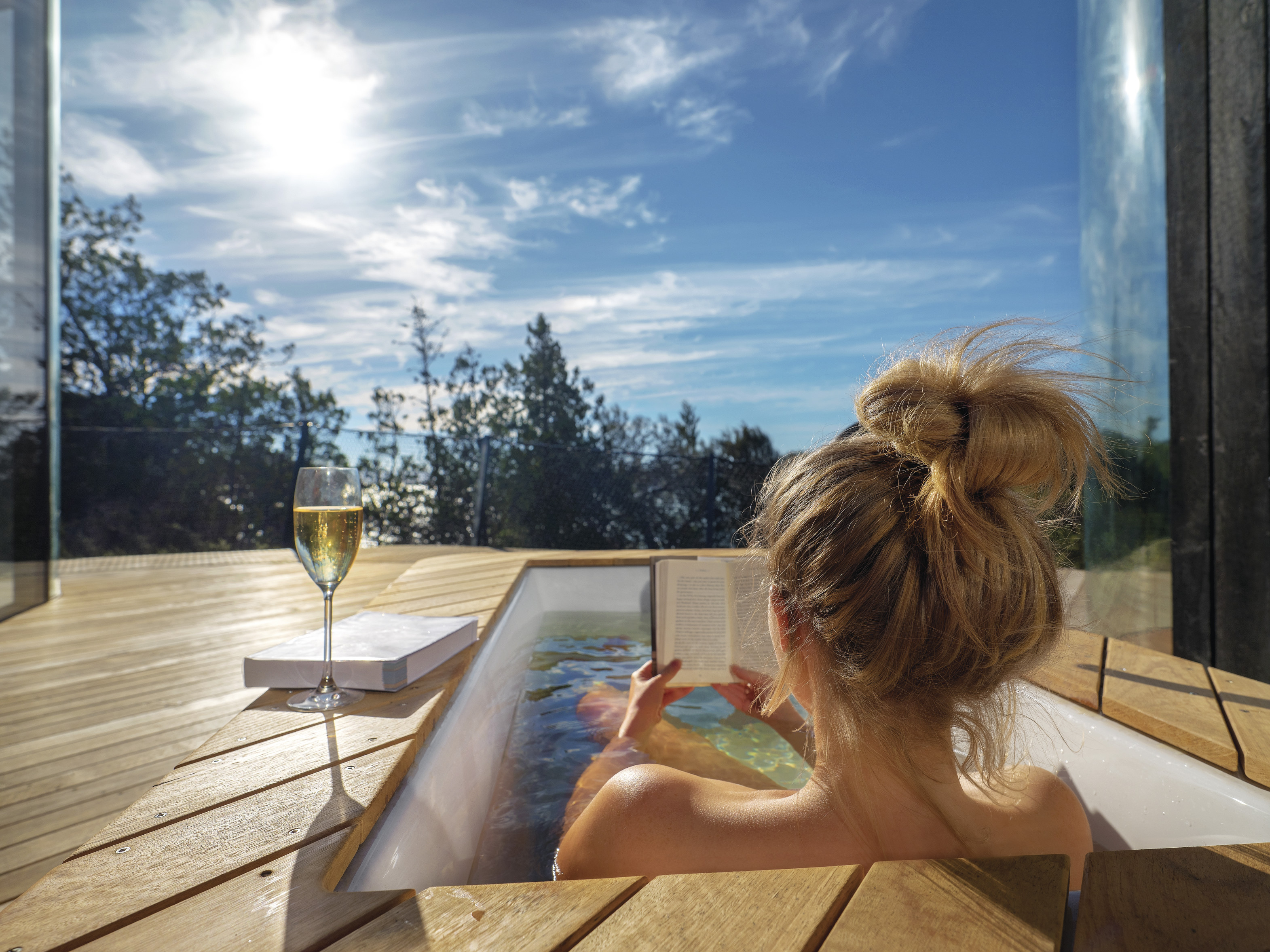 Woman relaxes reading a book with a glass of white wine in an outdoor bath tub at Freycinet Lodge, Coastal Pavilion accomodation. 