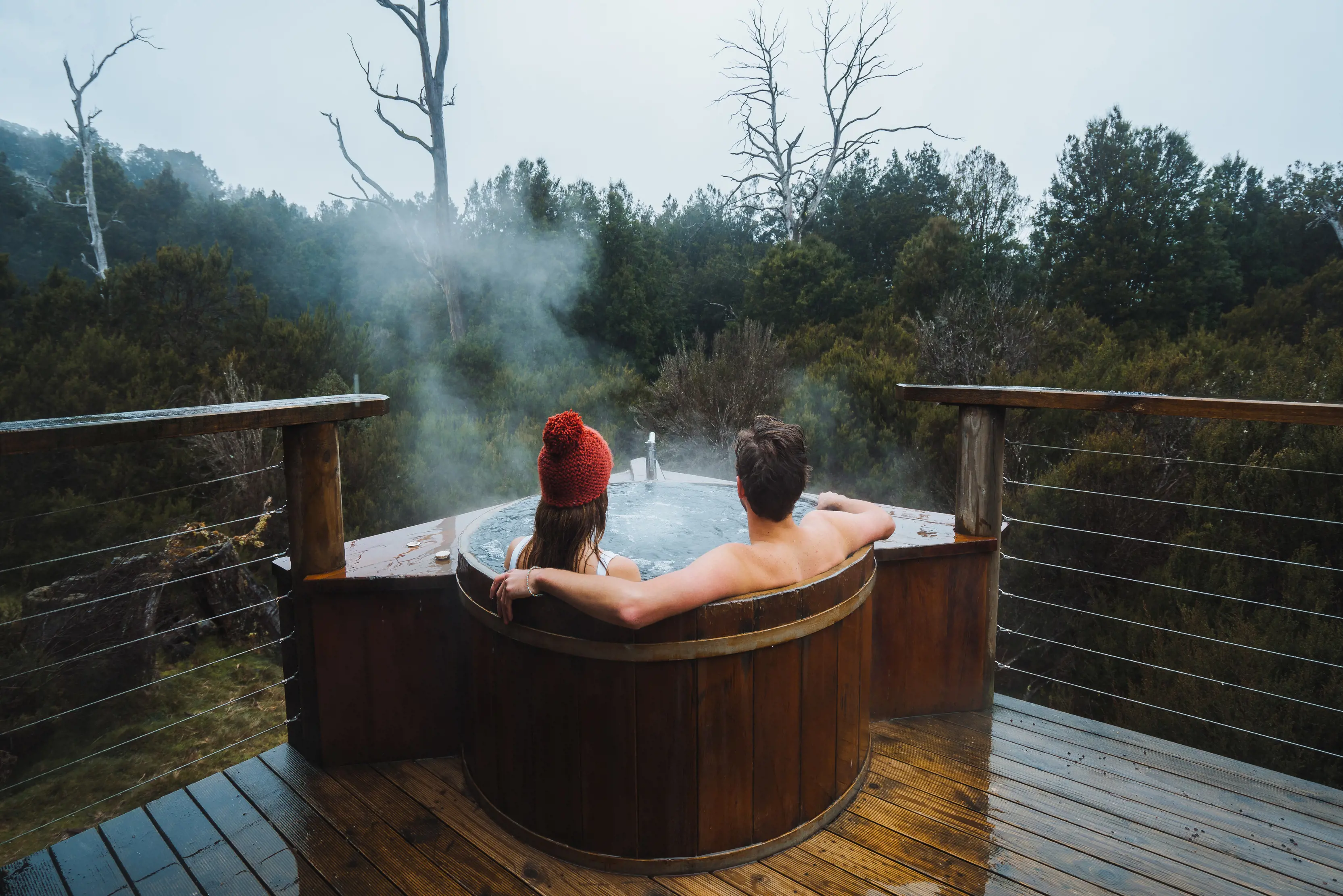 Couple relax in the steaming outdoor bath tub during winter at King Billy Suite, Peppers Cradle Mountain Lodge. The woman wears a read beanie.
