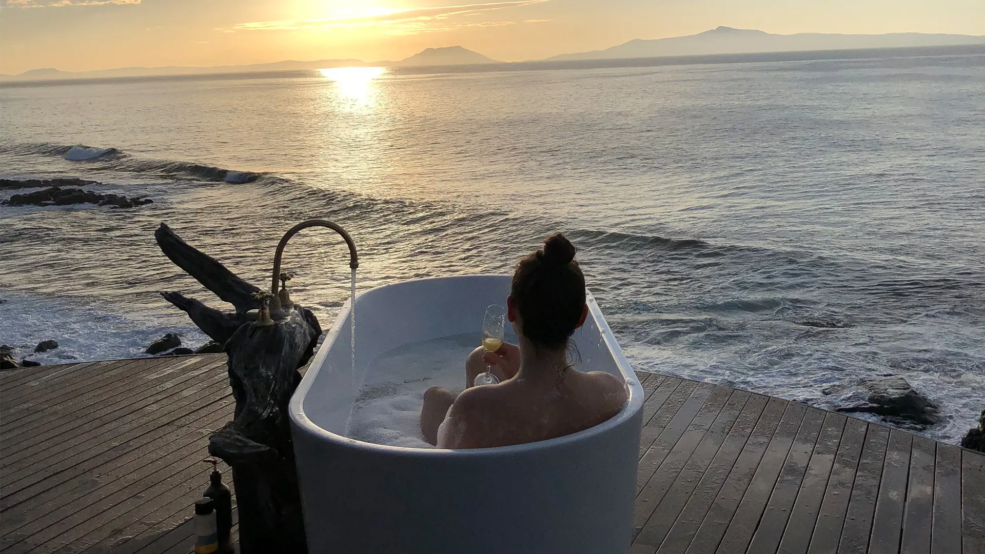 A woman sits in a white modern bath tub sitting on a wooden platform overlooking the beach and ocean at sunset.