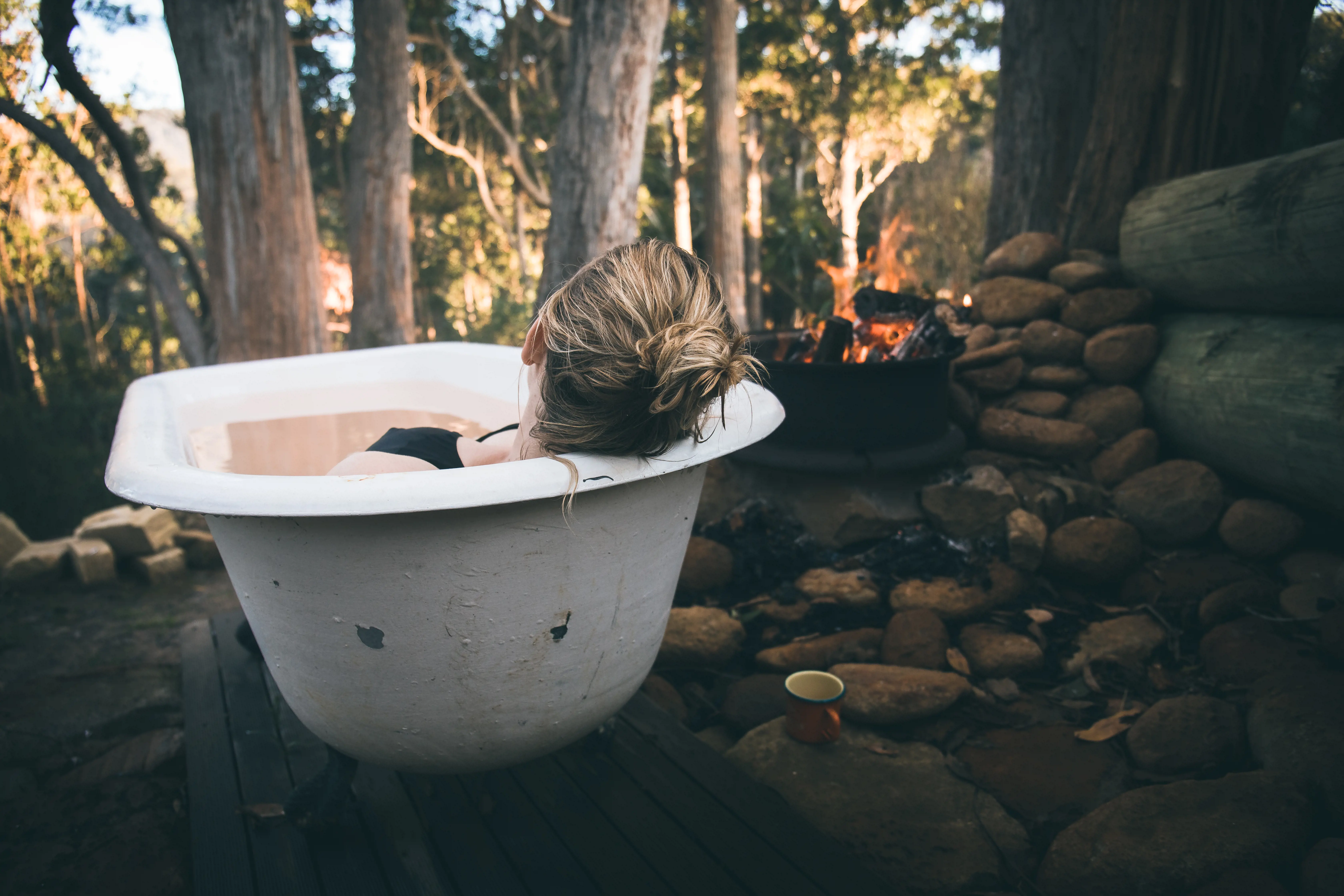 A woman reclines in a bath next to a wood fire in a eucalyptus forest.