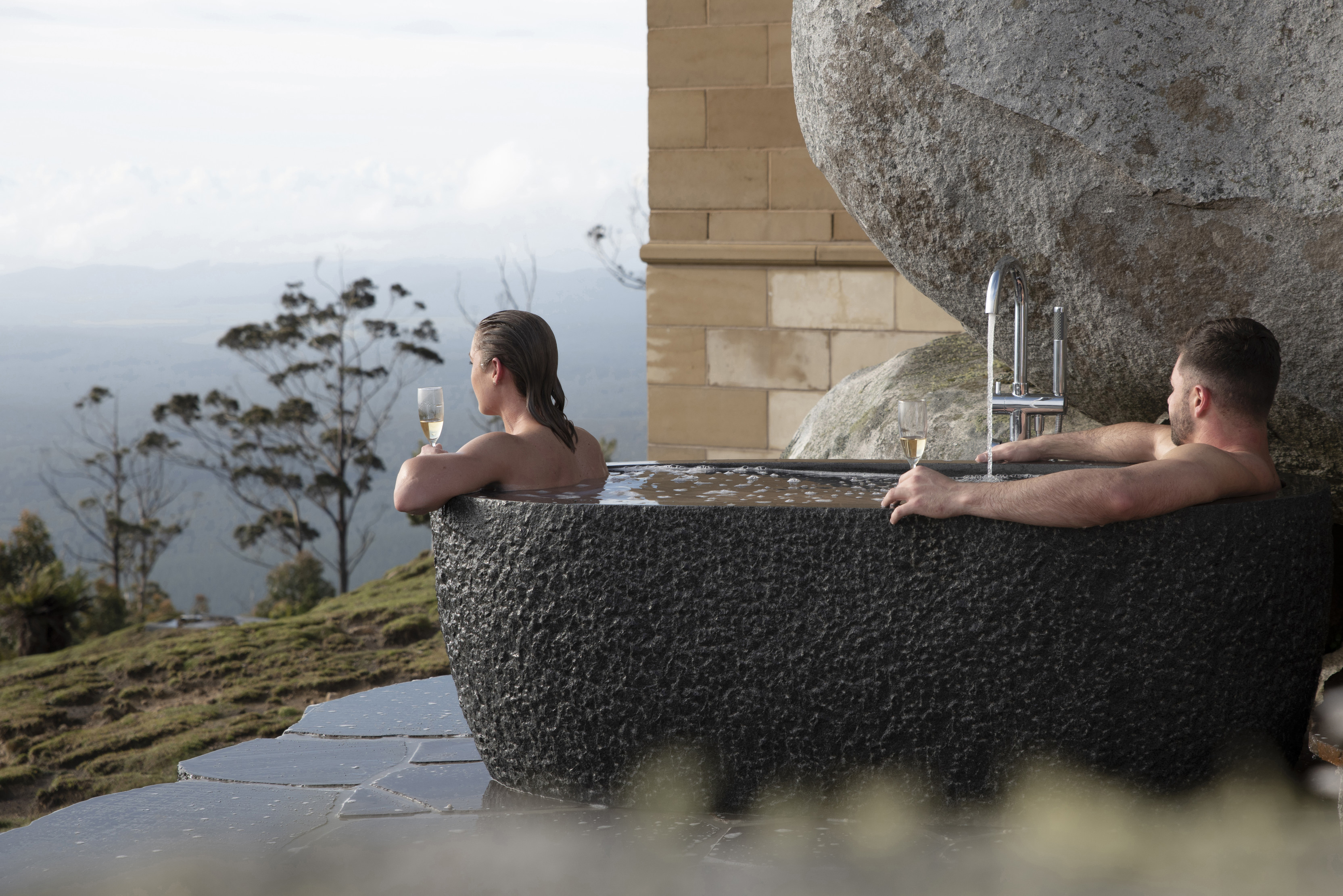 A couple relax with champagne in a hot outdoor bath at The Keep, perched upon a 650 metre rocky pinnacle overlooking Tasmania’s North east. 