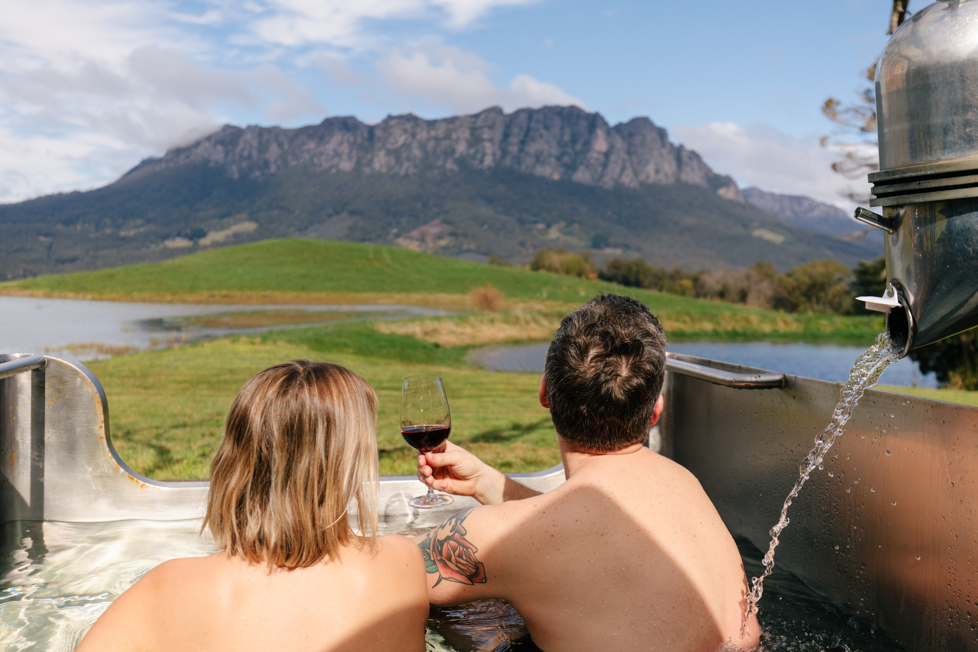 A couple relax with red wine in an outdoor bathtub at Eagles Nest Retreat. There are mountain views in the background. 