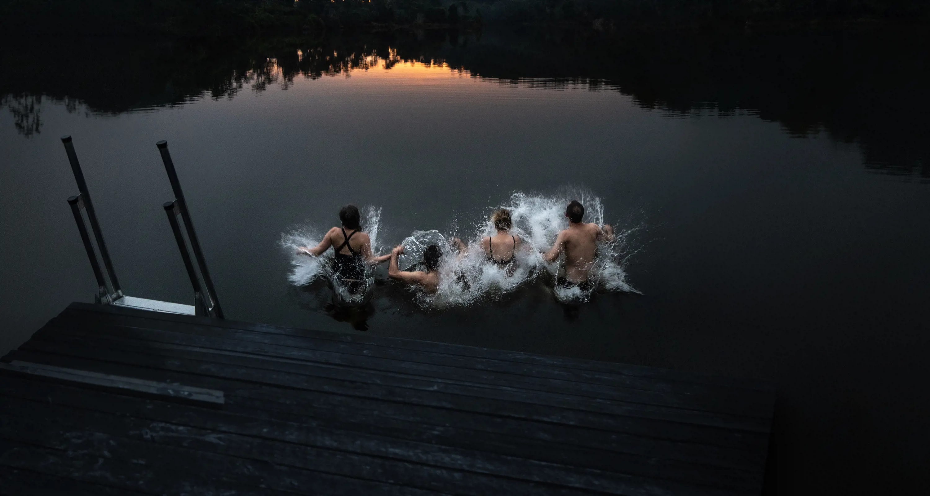 A group of four people jump into the dark waters of a lake at dusk.