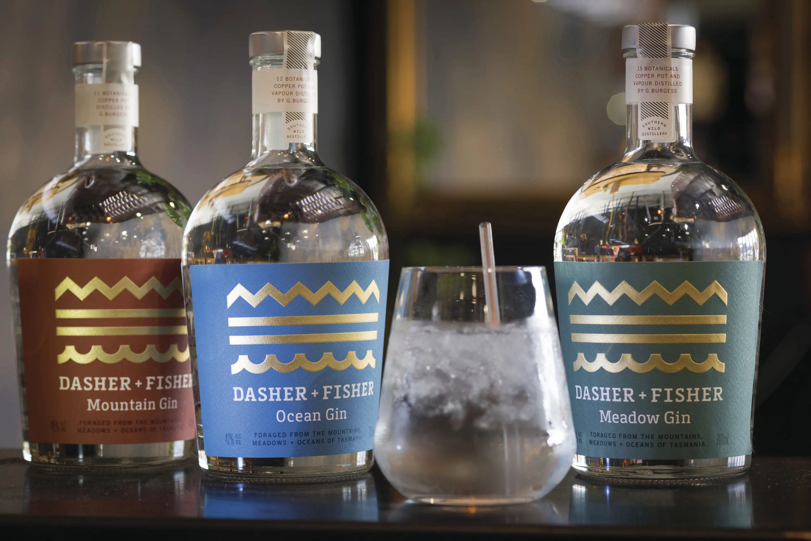 Three bottles of Dasher + Fisher Gin and a glass with drink poured at Southern Wild Distillery. The bottle labels include; Ocean, Meadow and Mountain Gin. 
