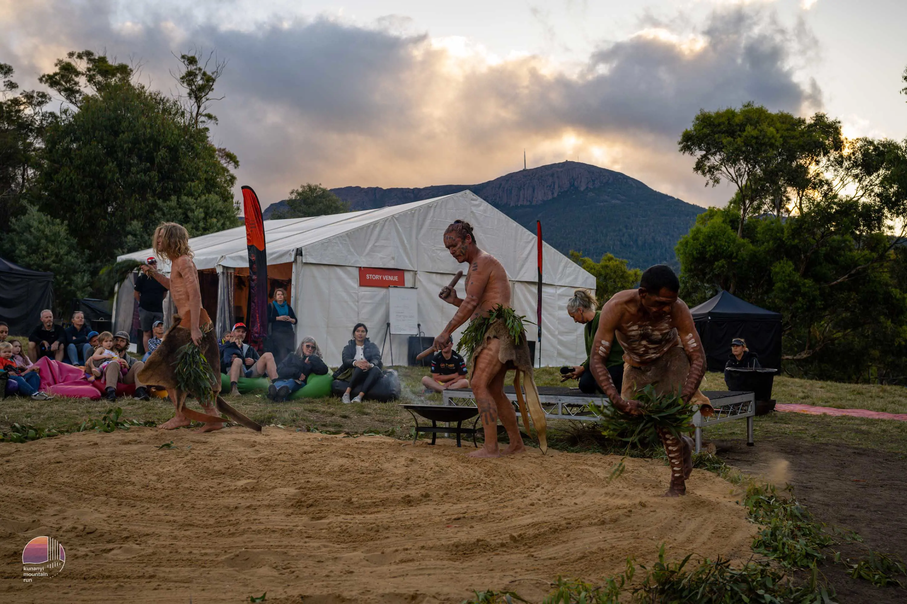 A group of Tasmanian Aboriginal men perform a ceremonial dance on an area prepared in front of tents for a running race.