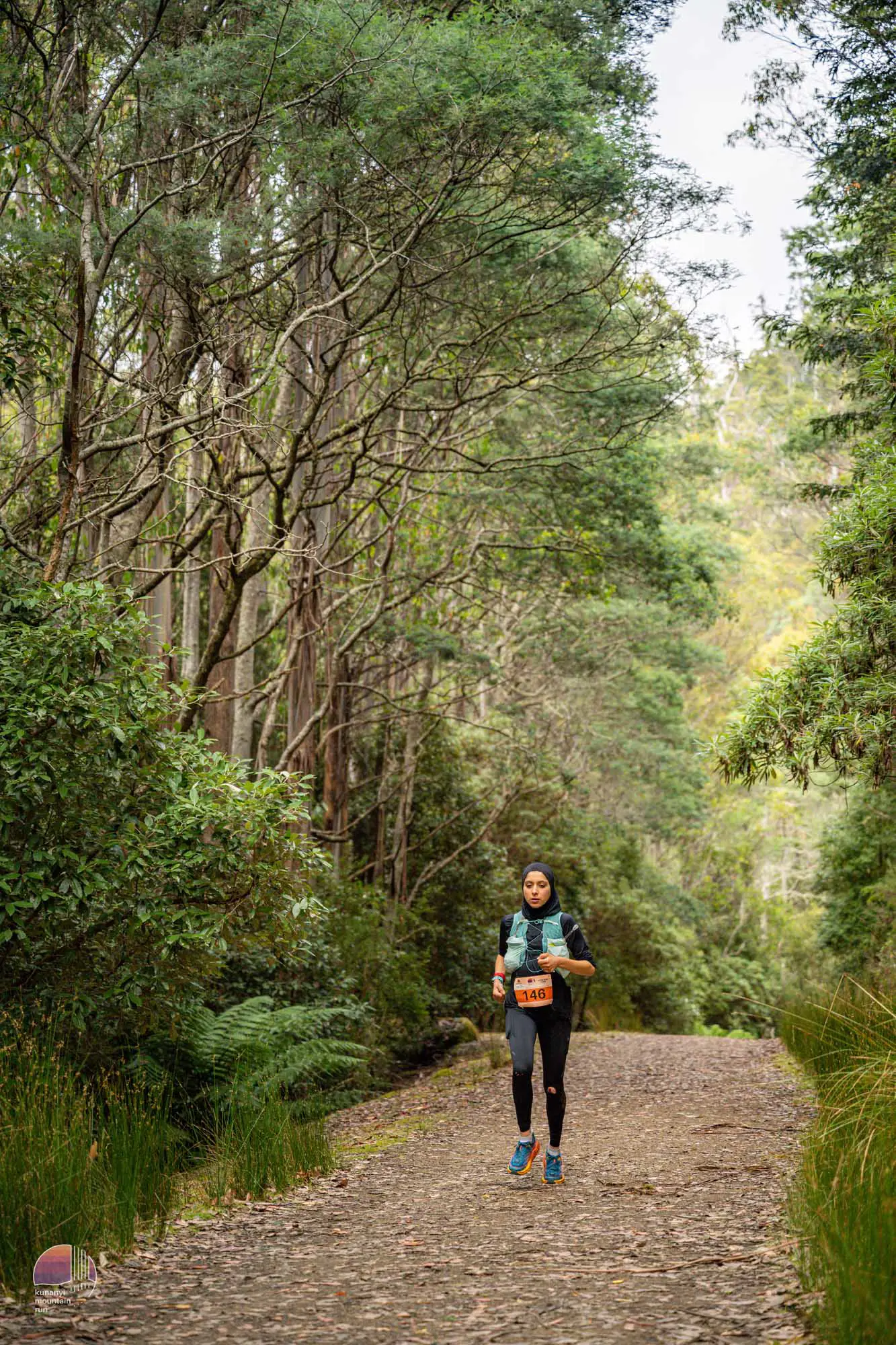 A young woman in a trail running vest, athletic gear and a hijab runs along a leafy path through dense eucalyptus forest. 