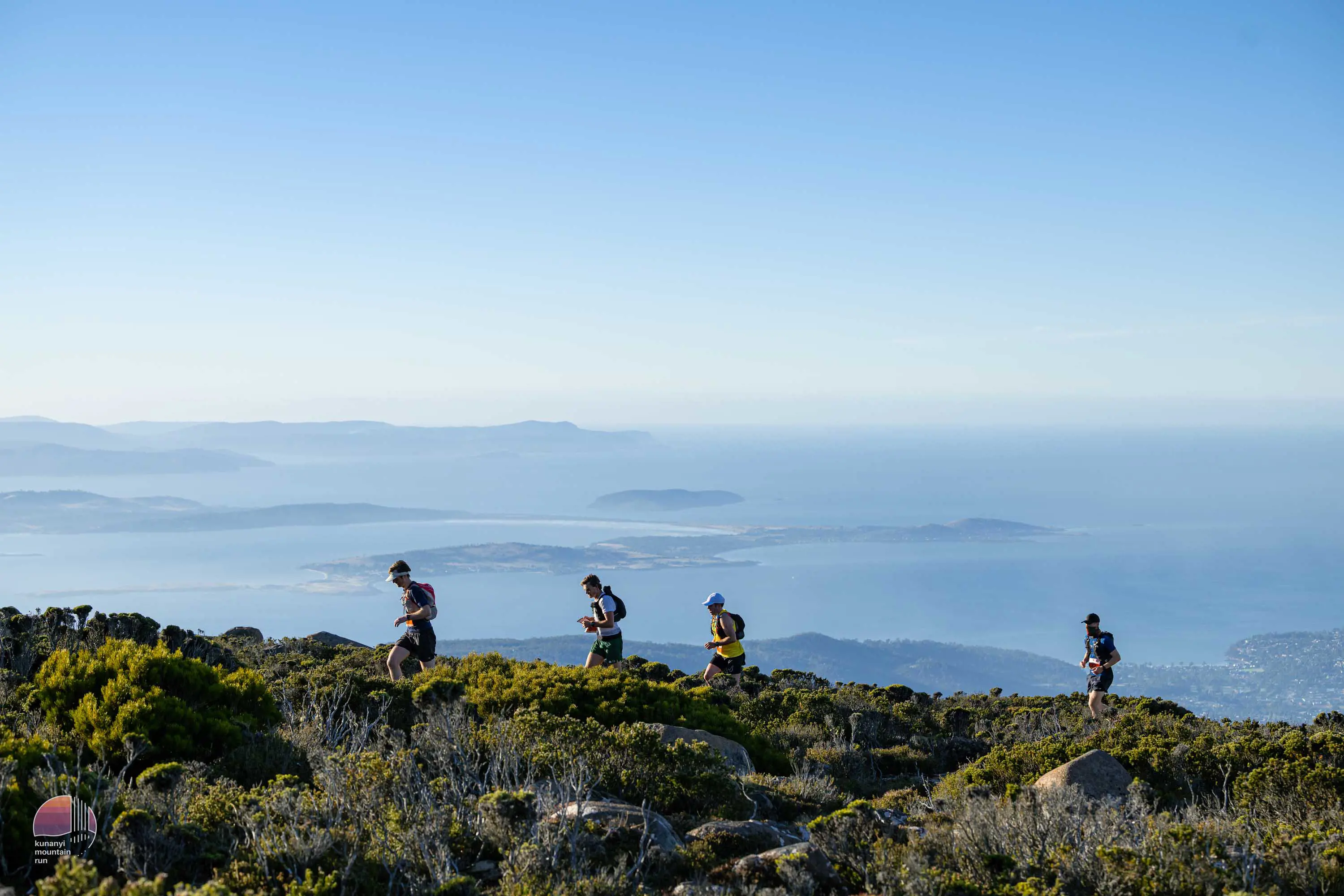 A group of four runners in hats, trail running vests and athletic gear run along an elevated ridge of low scrub with views of a cloud covered valley stretching into the distance.