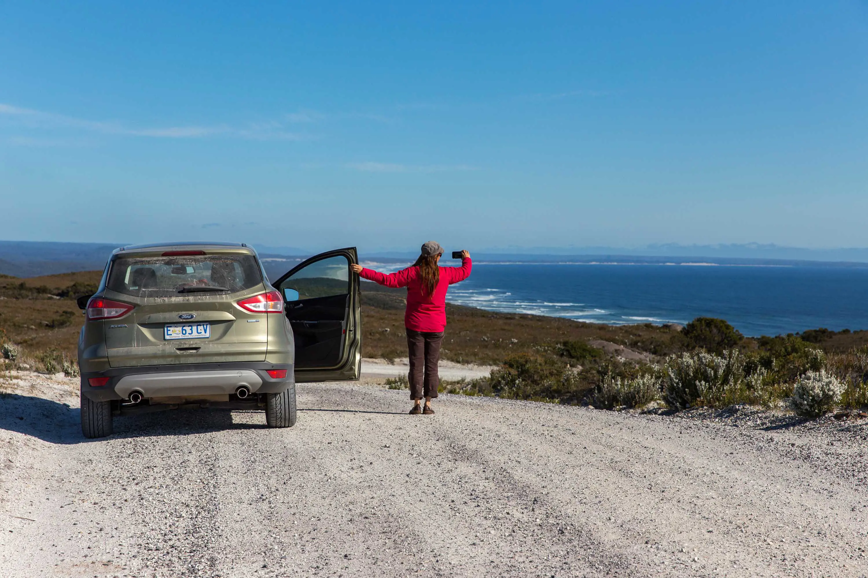 A woman wearing a red jacket stands beside a khaki coloured sport utility vehicle on a gravel road and looks at the ocean in the distance on a clear day. 