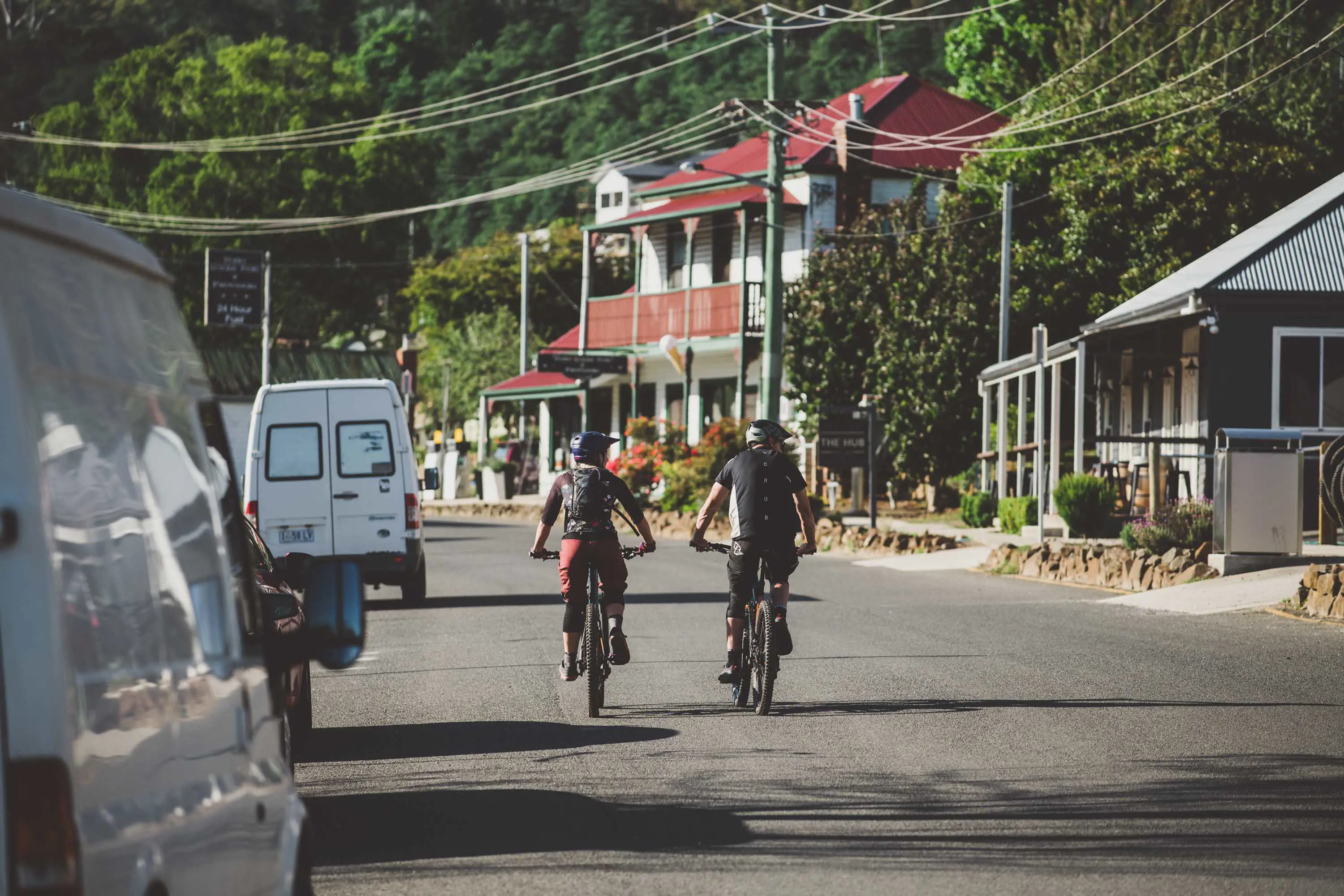 Two mountain bike riders ride their bikes along bitumen throuhg a  country town.