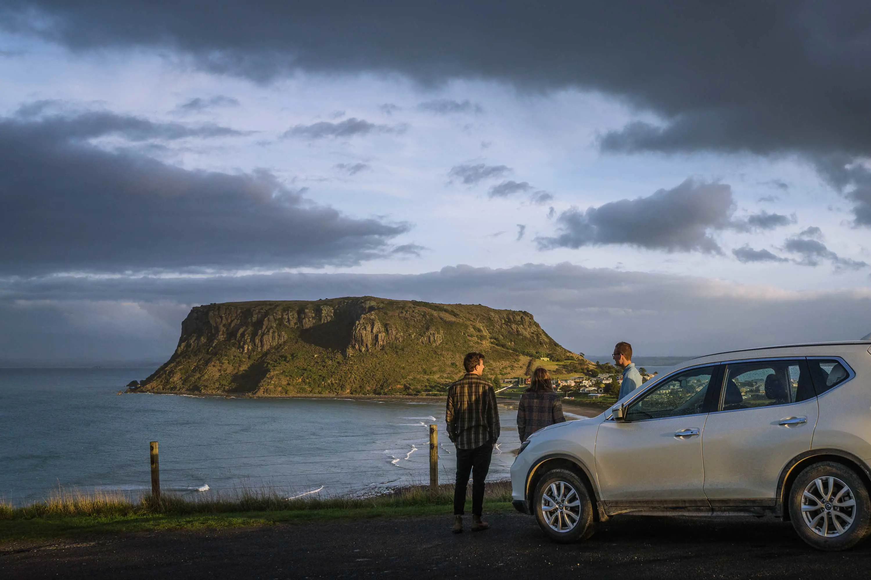 A group of three people stand in front of their silver sports utility vehicle and look out over a bay with a large, flat-topped peninsula in the background.