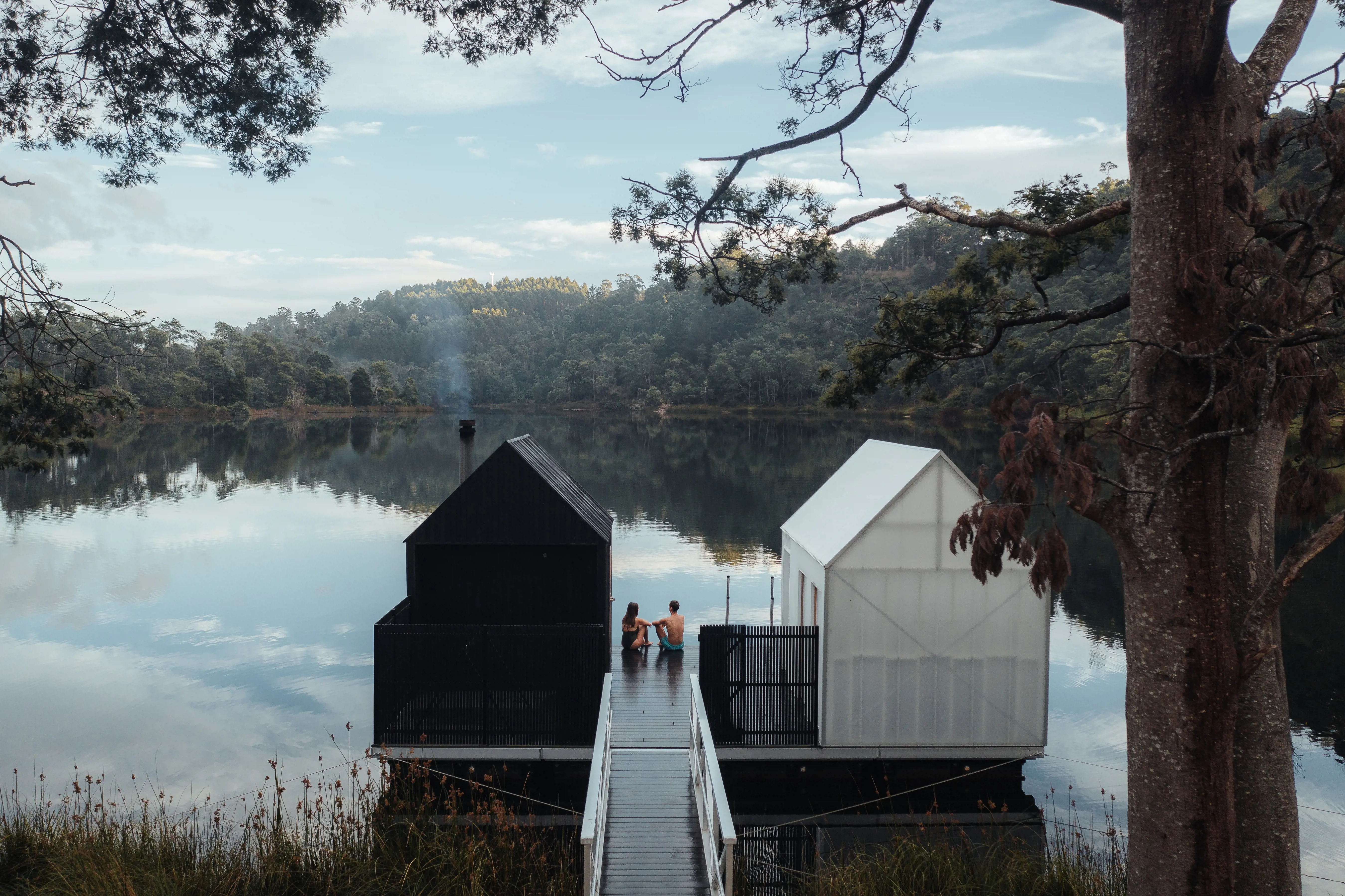 Bathers sitting on jetty between black and white sauna huts, overlooking lake.