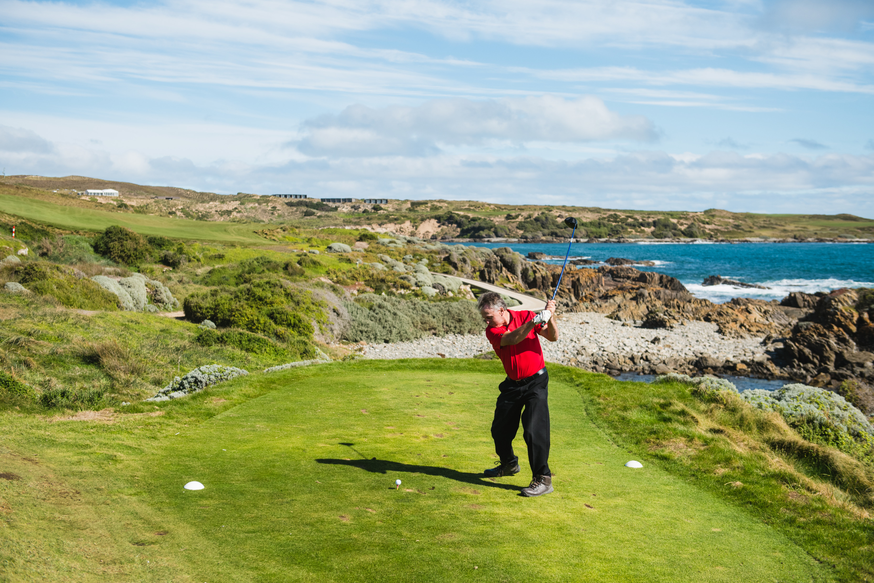 A man swings his golf club at Cape Wickham Golf Course, with lush green landscapes and ocean in the background.