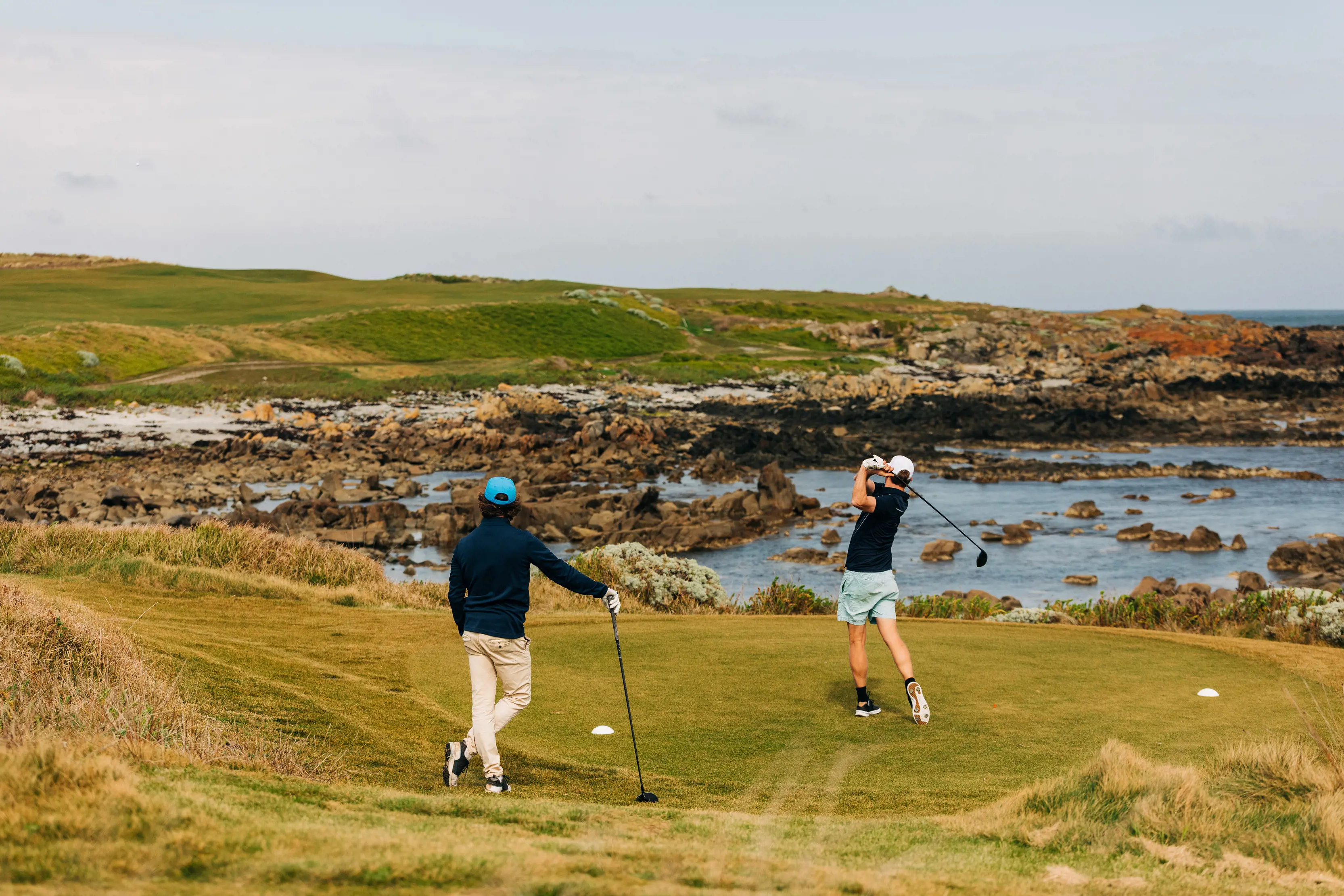 A golfer watches his friend take a swing at Ocean Dunes Golf Course, King Island. Landscape views in the background.