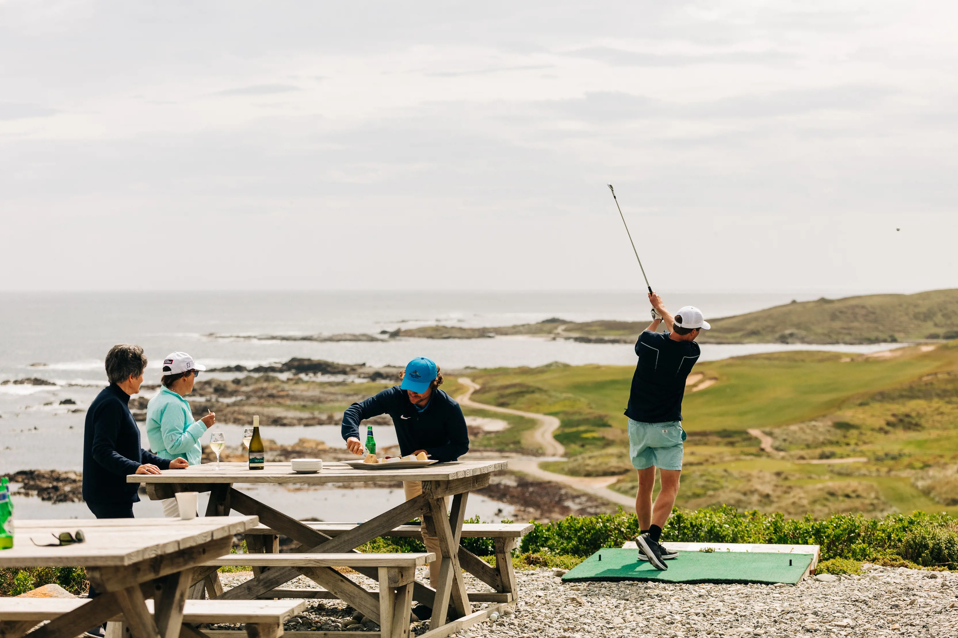 A group of friends enjoy food and drink on an outside bench whilst another practices their shot at Ocean Dunes Golf Course, King Island.