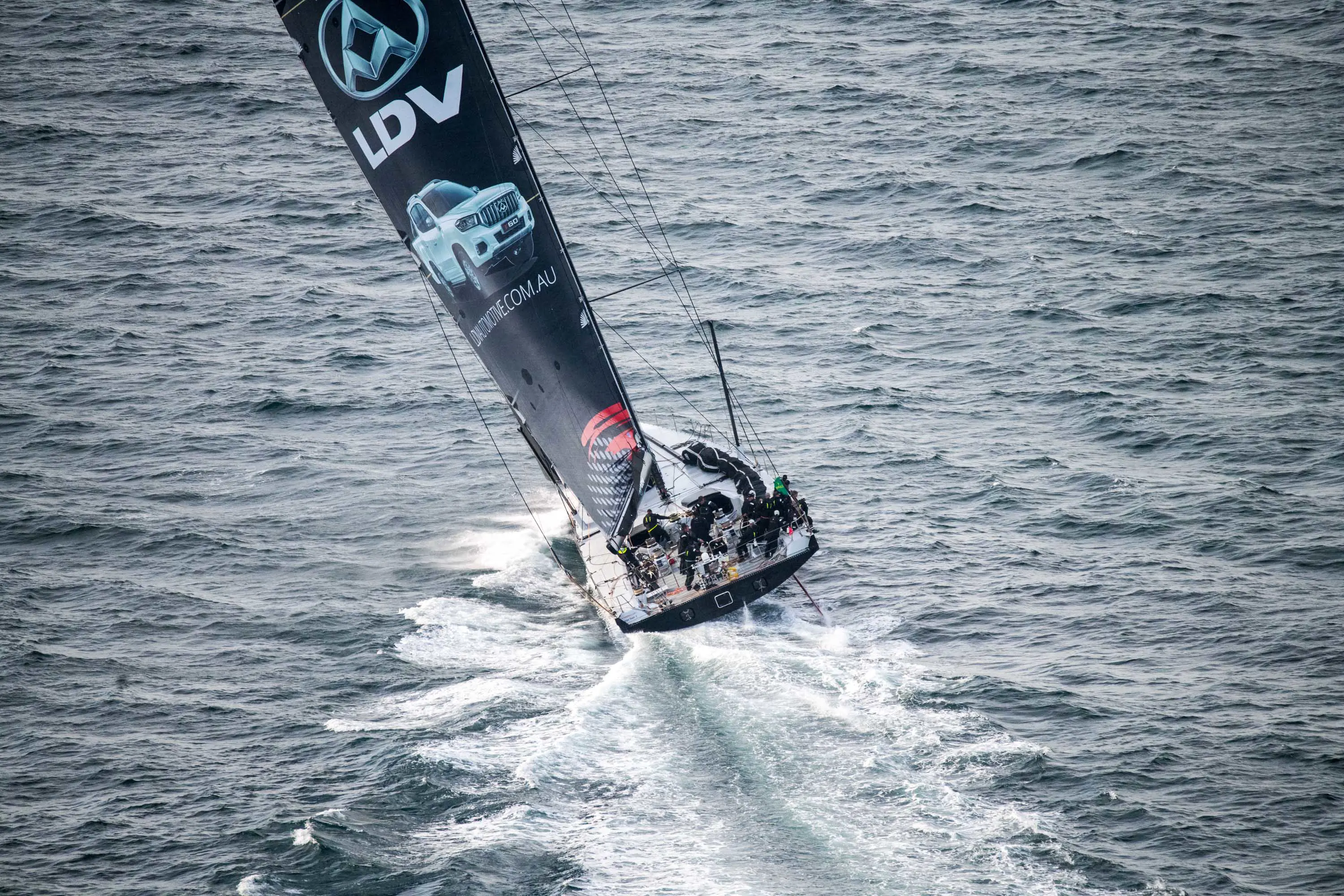 A racing yacht with a large black sail leans in to a turn on wavey seas.