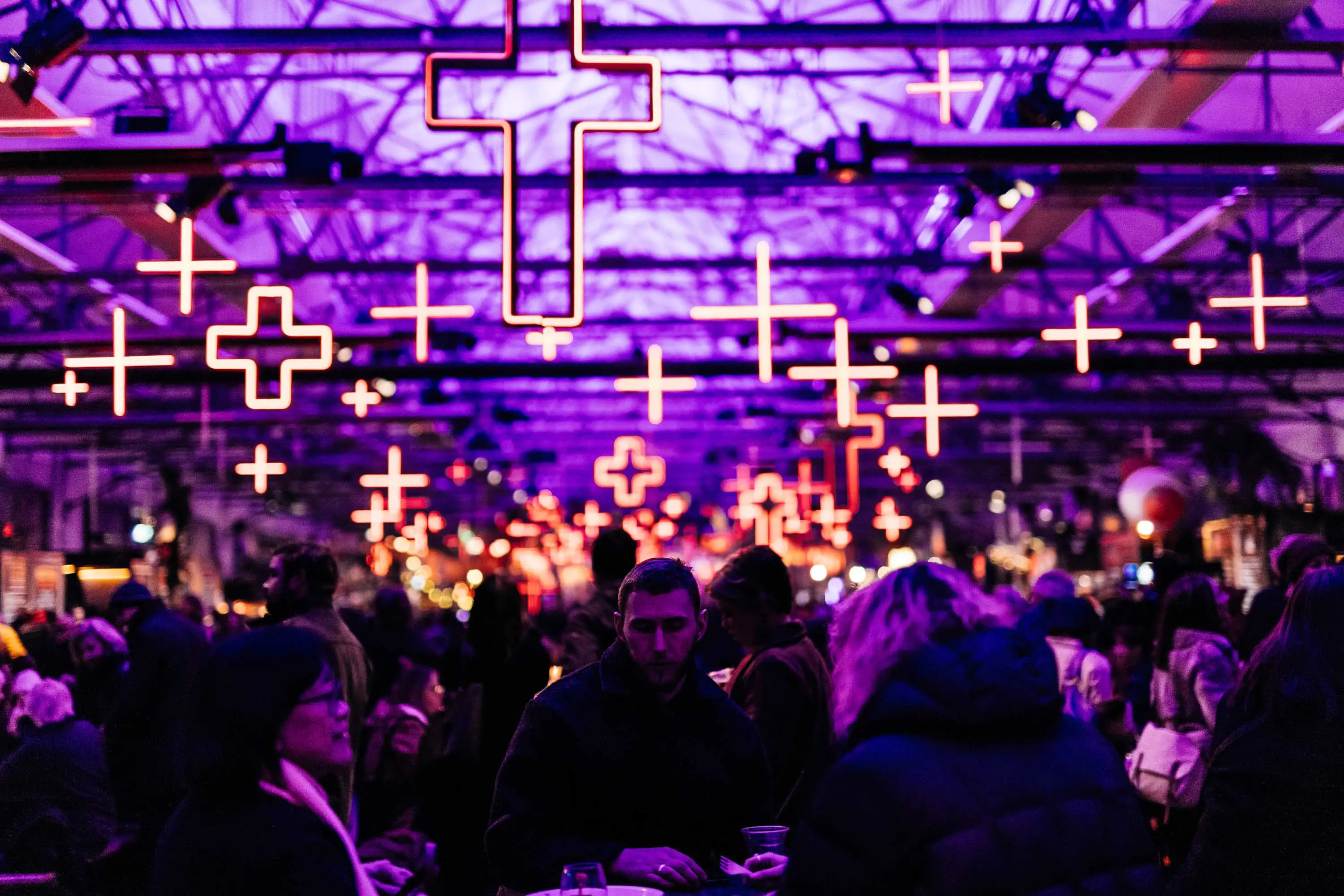A large group of people sit at tables under moody purple and yellow lighting in a high-ceiling wharf shed.