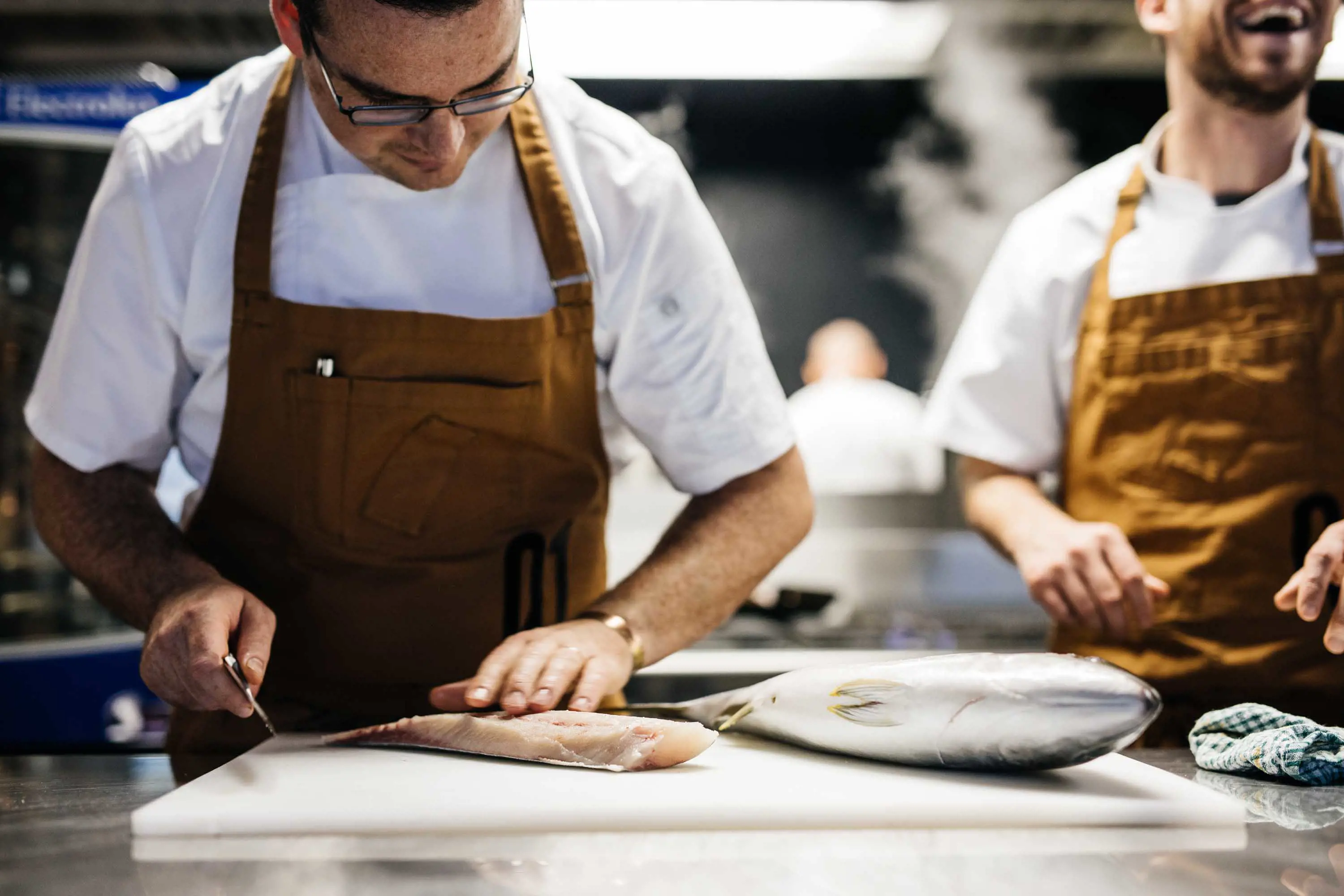 Two chefs work in the stainless steel kitchen. One slices fresh fish on a white plastic cutting board. 