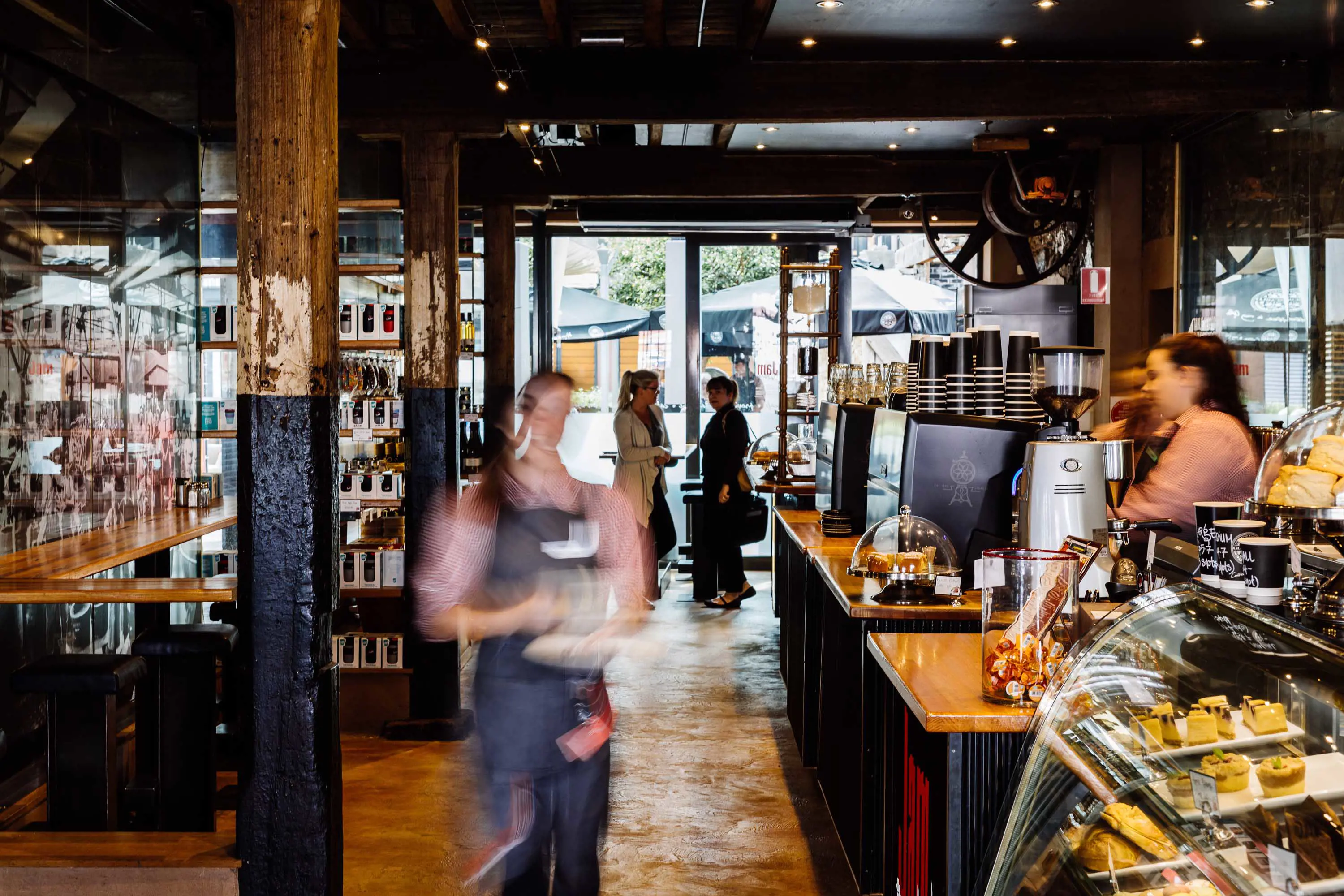 A cafe with glass cabinets and wooden beams is bustling with staff and customers.